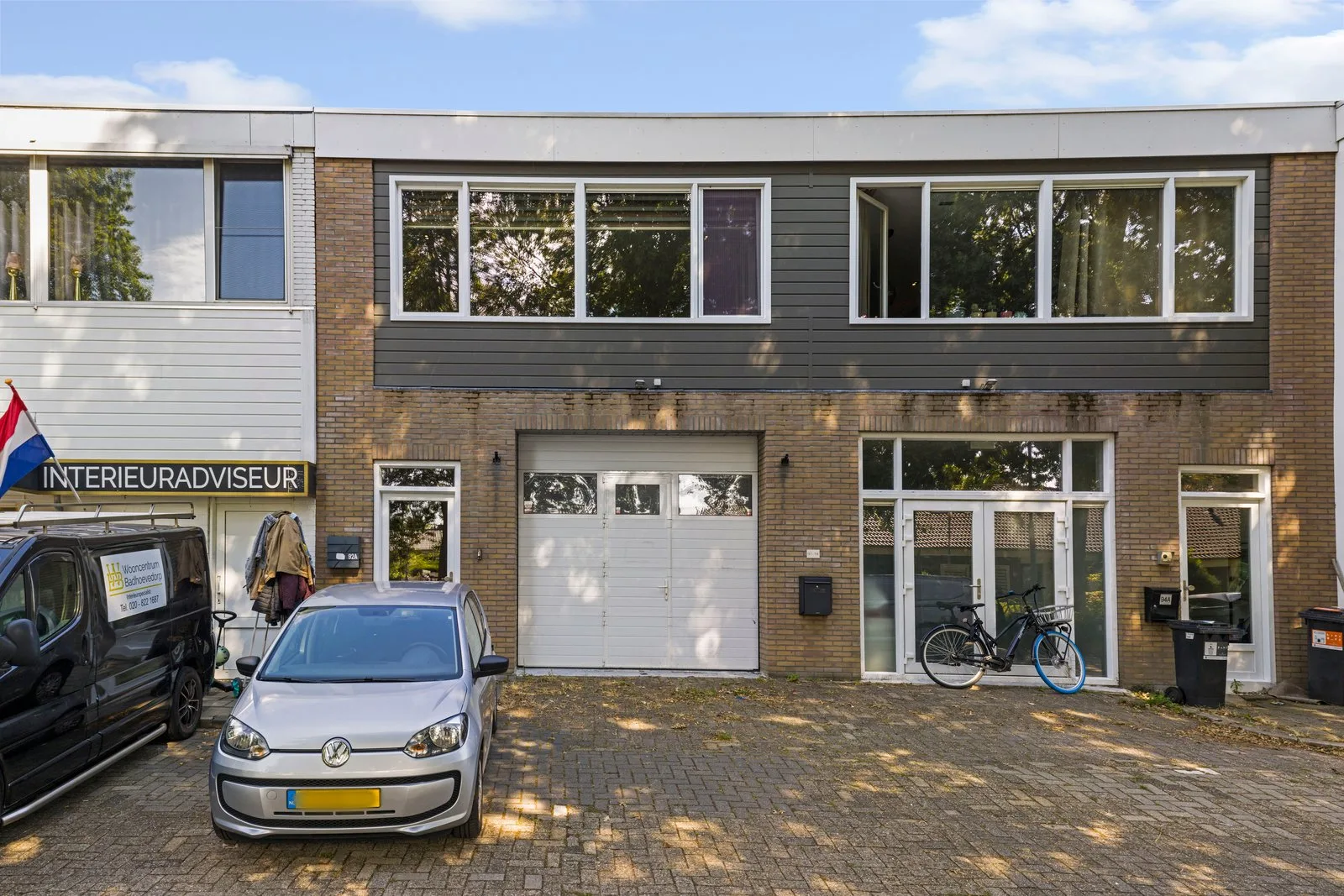 Bakstenen gevel met donkergrijze bekleding op de verdieping, witte garagedeur en een bestraat voorterrein