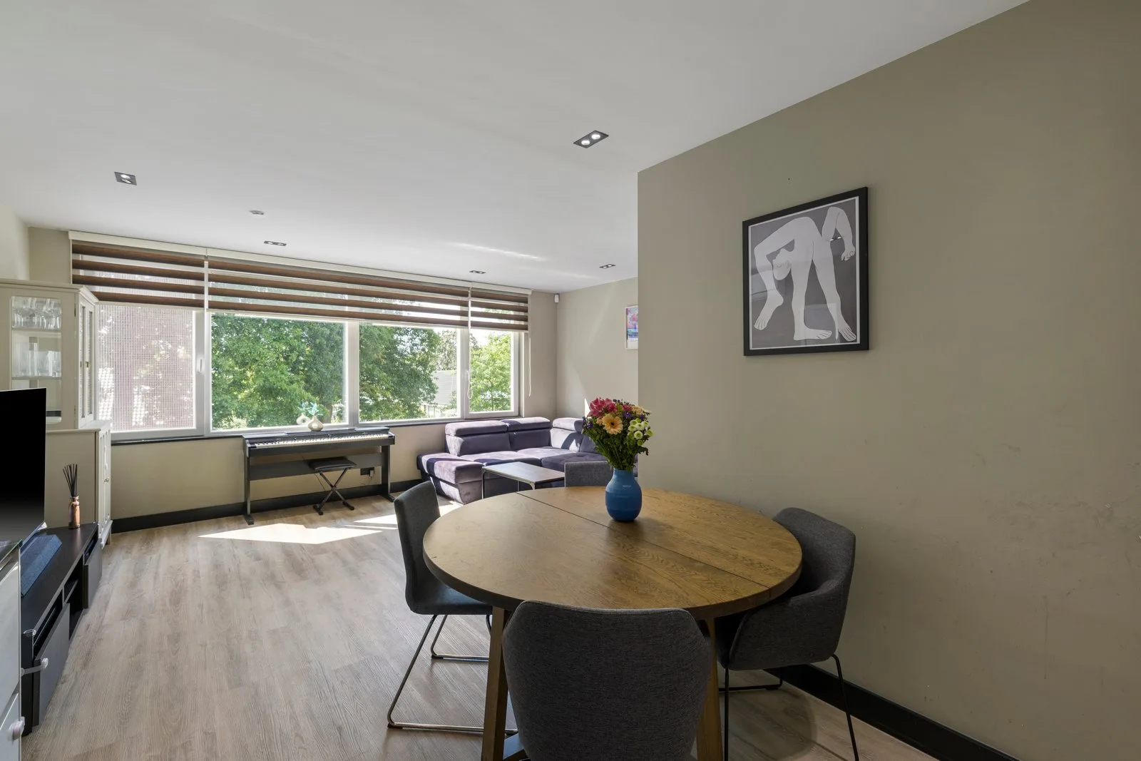 Ronde houten eettafel met grijze stoelen, daarachter de zithoek en een brede raampartij met bomen