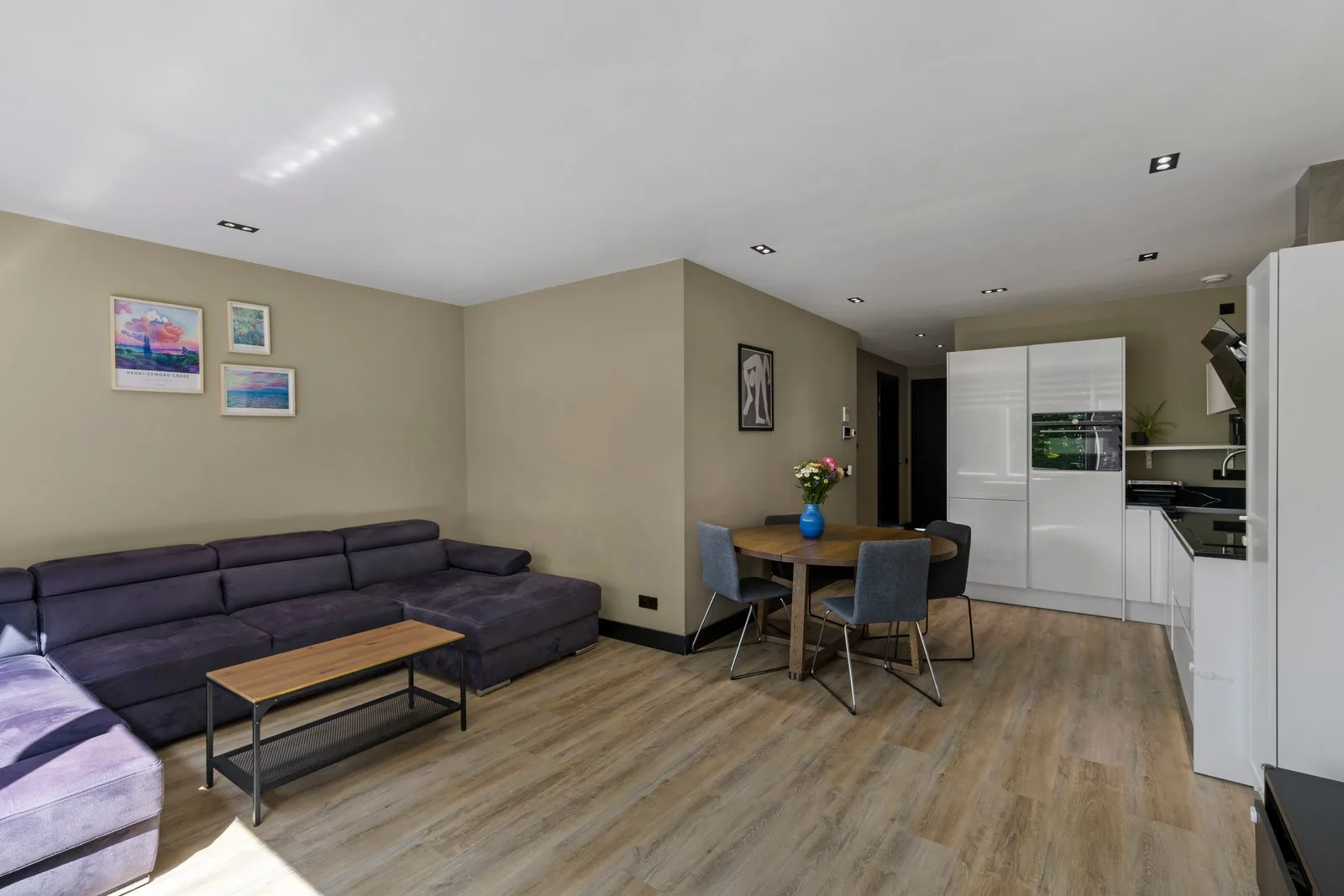 Woonkamer gezien vanuit de zithoek, over de lichte vloer naar de eettafel en de witte keuken