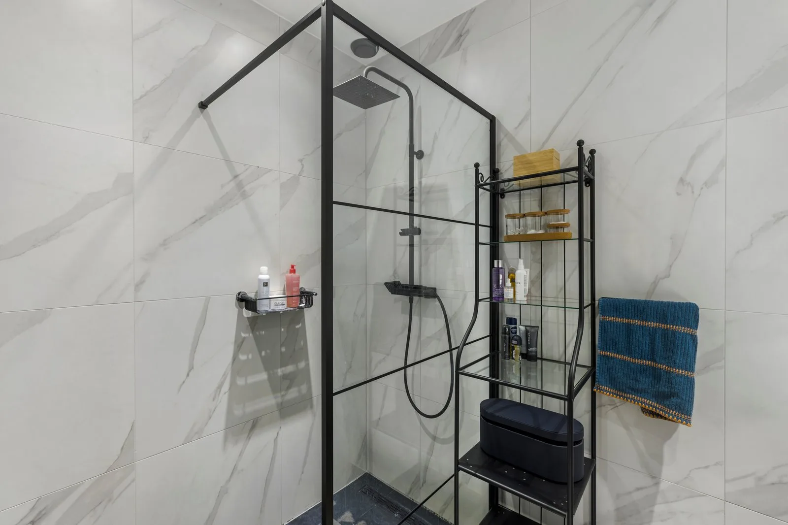 Badkamer met inloopdouche achter glas in een zwart frame, naast een zwarte stellingkast met handdoek