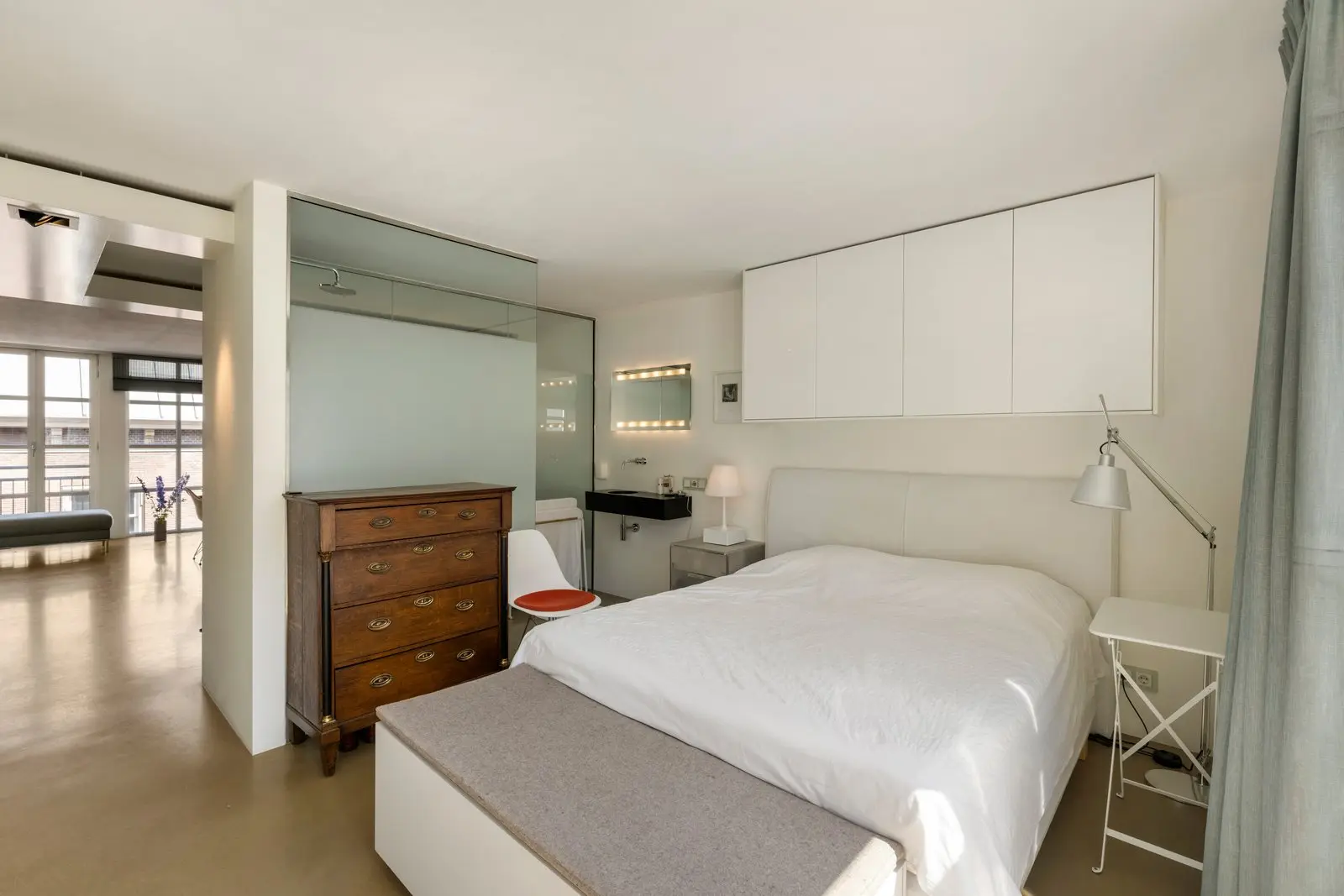 Tweepersoonsbed en houten ladekast in de slaapkamer, met links de wastafel en een doorkijk naar de woonkamer