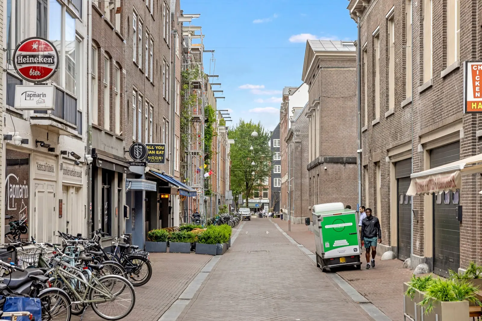 Straat met horecazaken, plantenbakken op de stoep, geparkeerde fietsen en een kleine groene bestelwagen