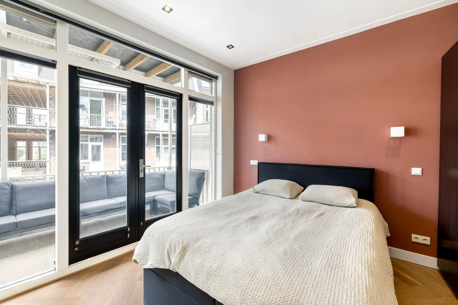 Slaapkamer met terracotta accentwand, donker bed en openslaande deuren naar een balkon met loungebank
