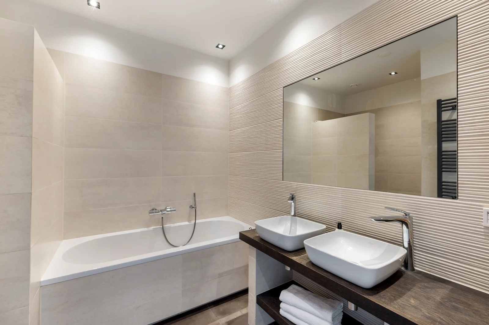 Badkamer met ligbad en twee witte wasbakken op donker hout, tegen geribbelde beige tegels