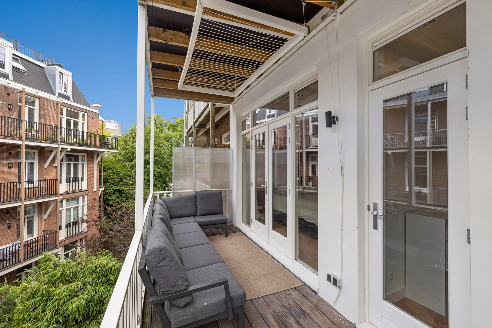 Grijze hoekbank met beige buitenkleed op het houten balkon, langs witte openslaande deuren met bovenlichten