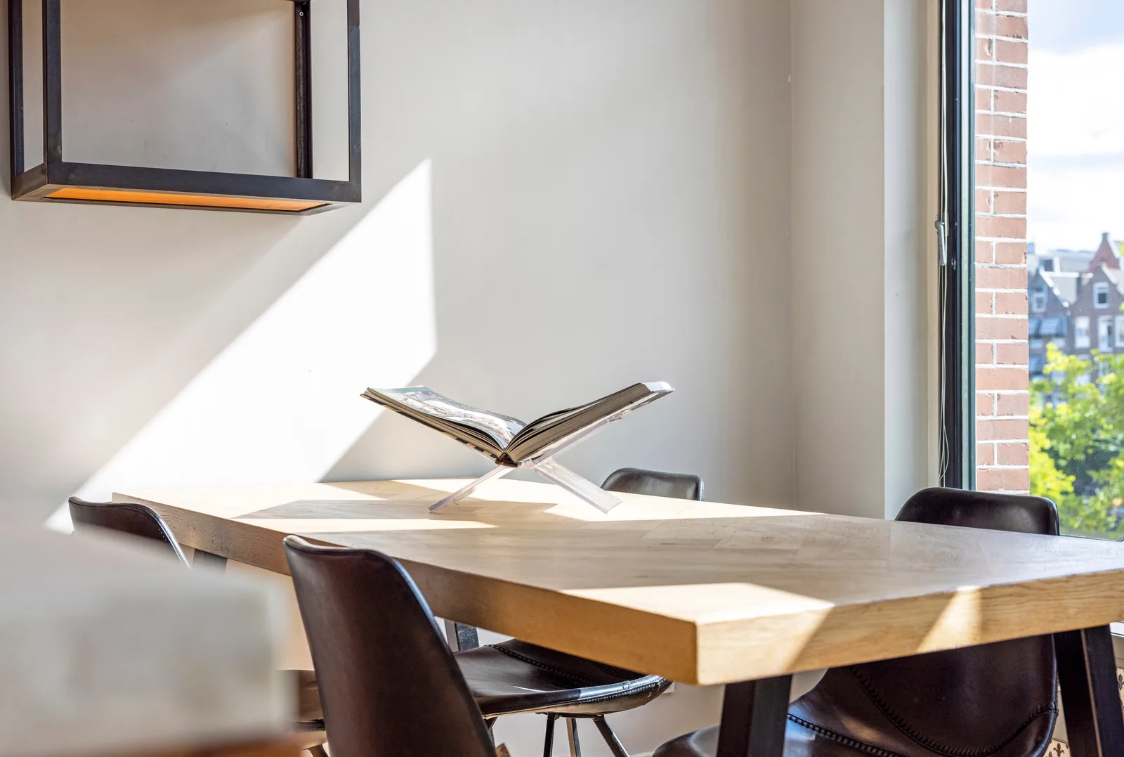 Lichthouten tafel met een opengeslagen boek op een doorzichtige standaard, eromheen donkerbruine leren stoelen