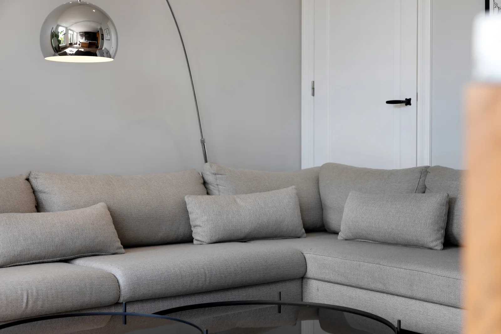 Lichtgrijze stoffen hoekbank met losse kussens onder een chromen booglamp, rechts een witte deur