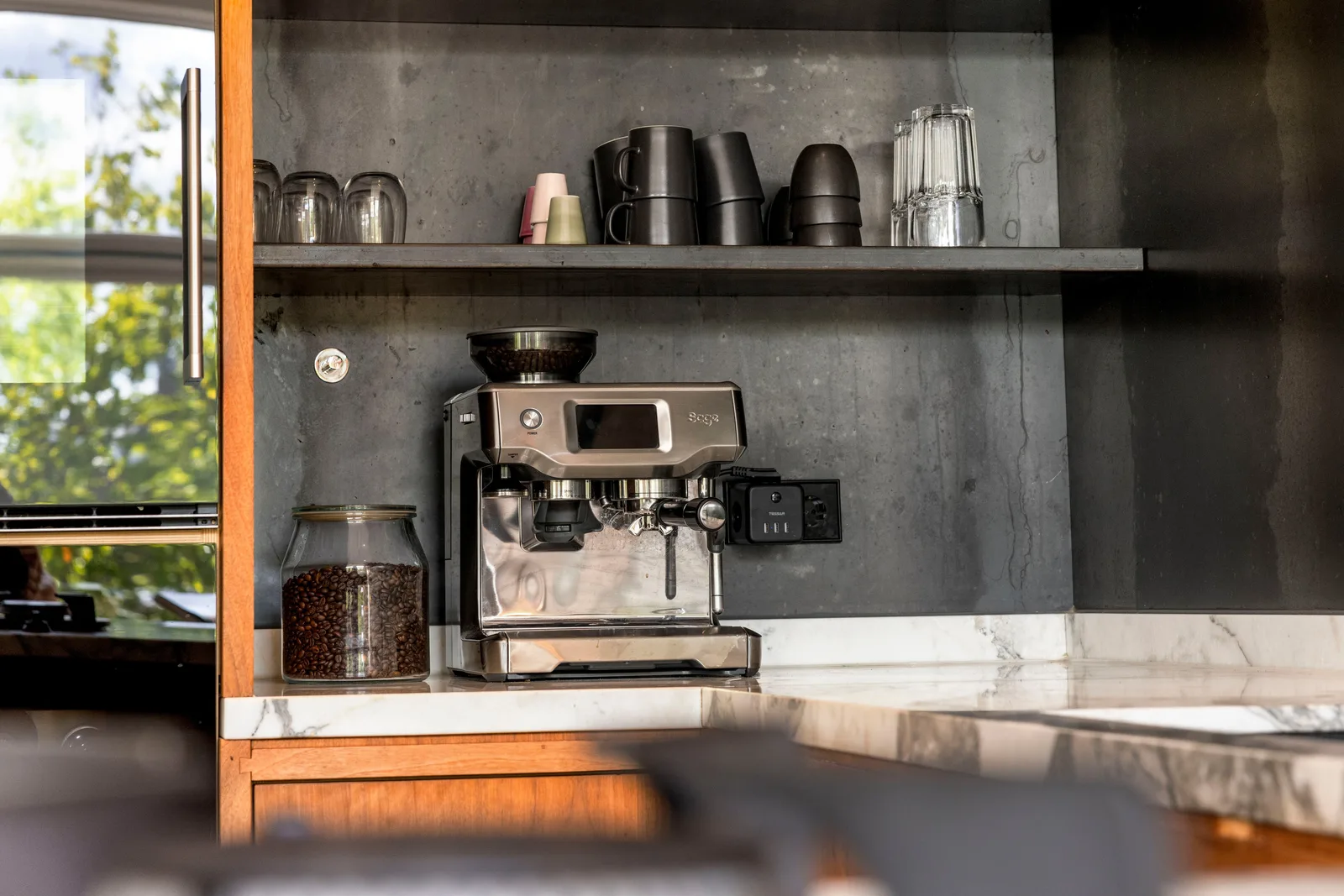 Espressomachine en een pot koffiebonen op marmer, daarboven een plank met mokken tegen een donkergrijze wand
