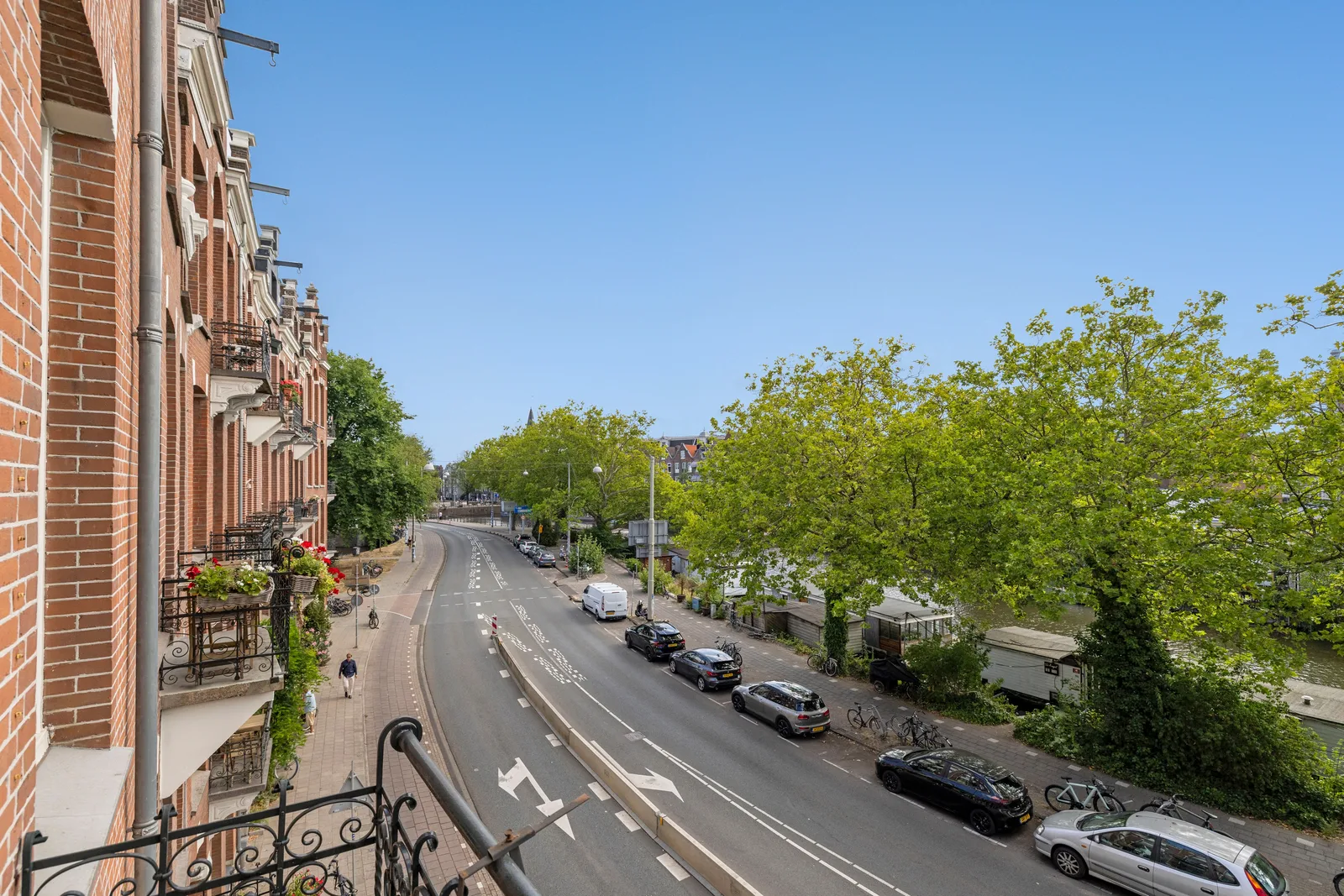 Over het smeedijzeren balkonhek naar de brede weg met geparkeerde auto's, bomen en water rechts
