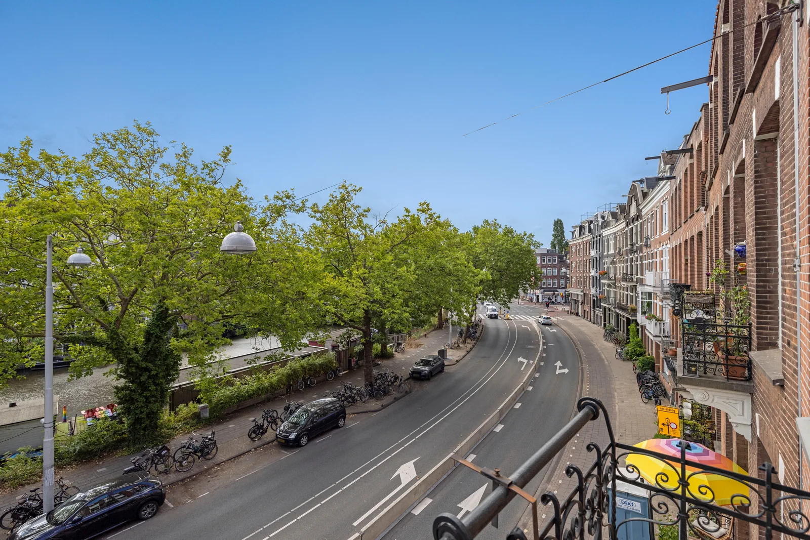 Over het smeedijzeren balkonhek zicht op de kade, links hoge bomen en water, rechts bakstenen huizen