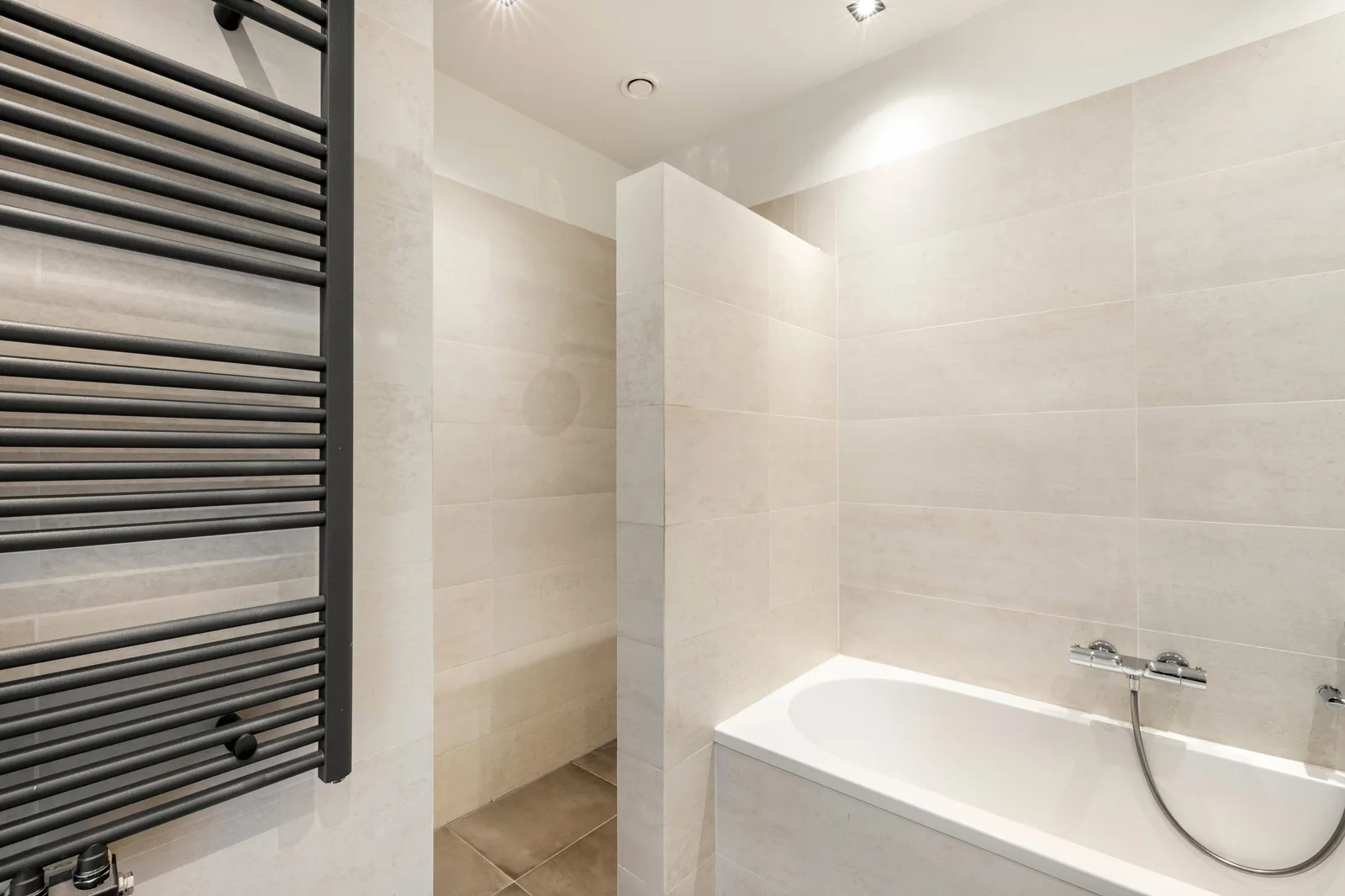 Beige betegelde badkamer met wit ligbad rechts, antracieten handdoekradiator links en een nis met grijsbruine vloertegels