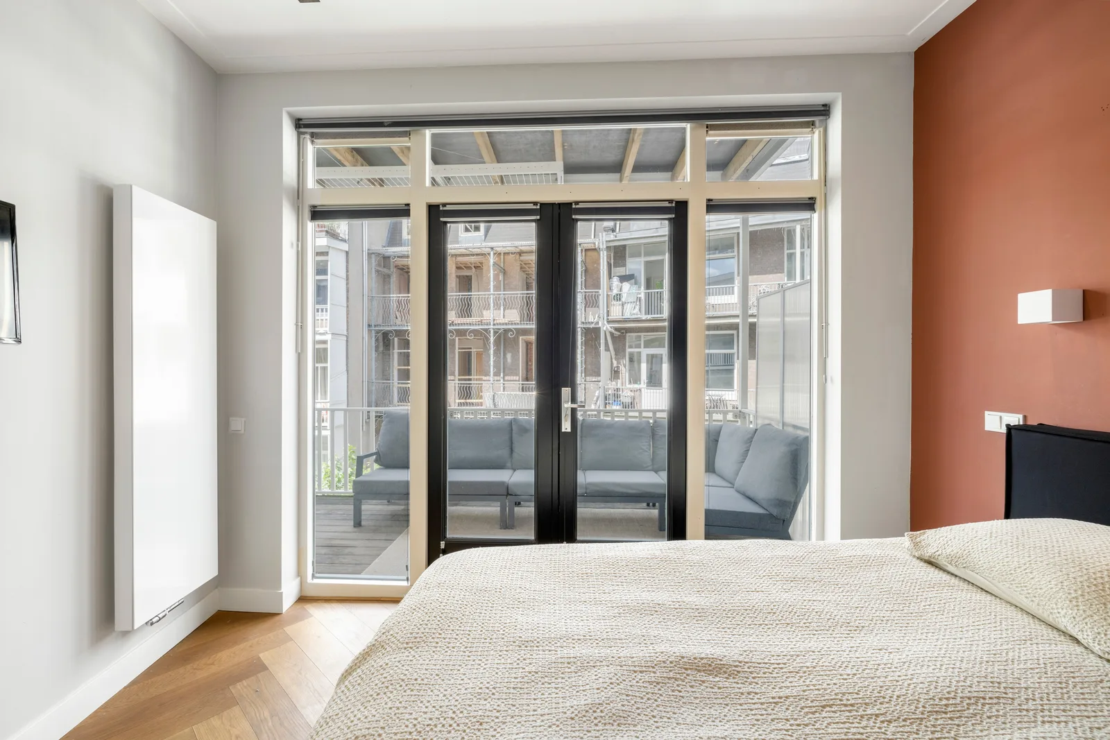 Bed tegen een terracotta wand, links een visgraatvloer en gesloten zwarte balkondeuren met grijze loungebank erachter
