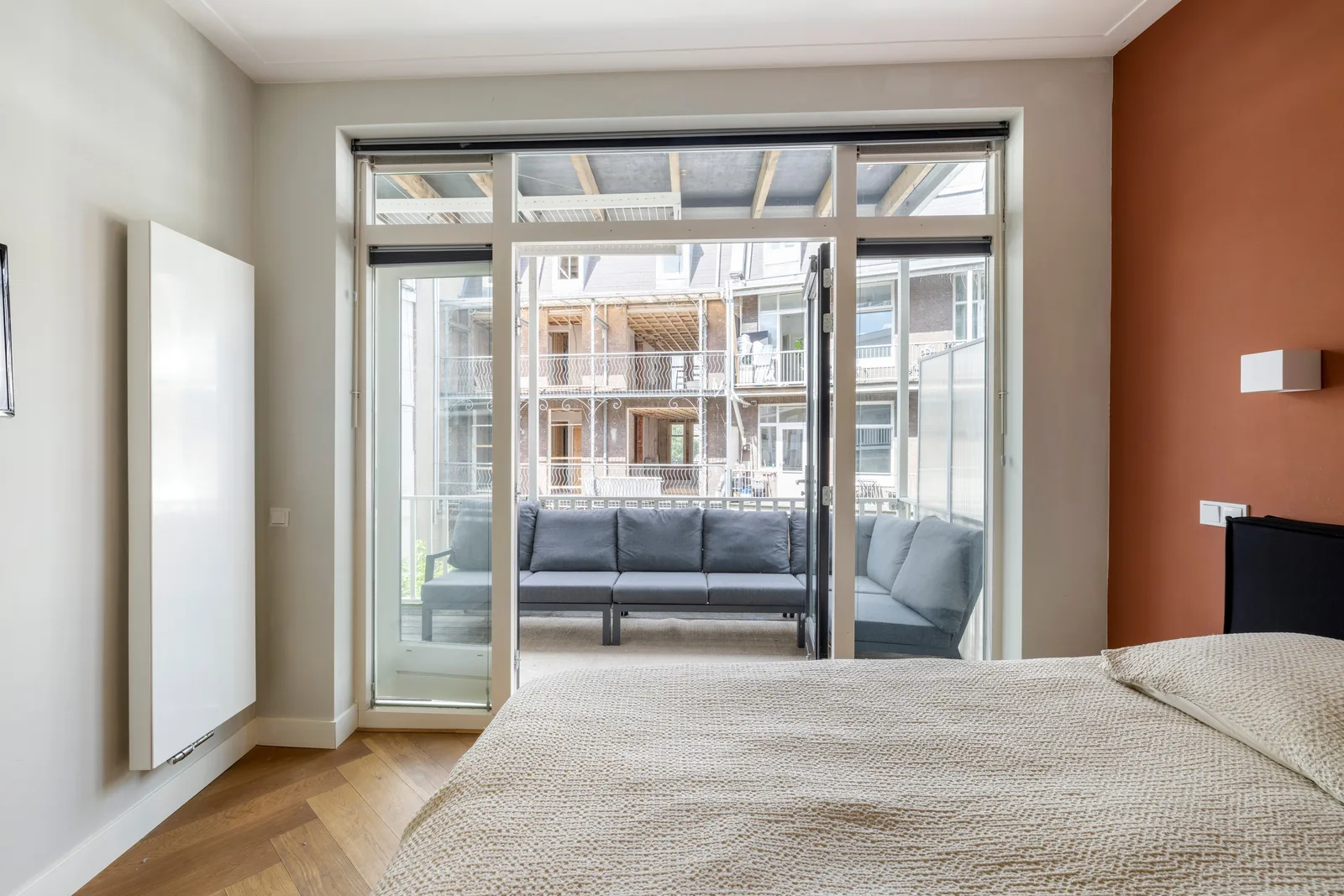 Bed met beige gebreide sprei tegen terracotta wand, openstaande deur naar balkon met grijze loungebank
