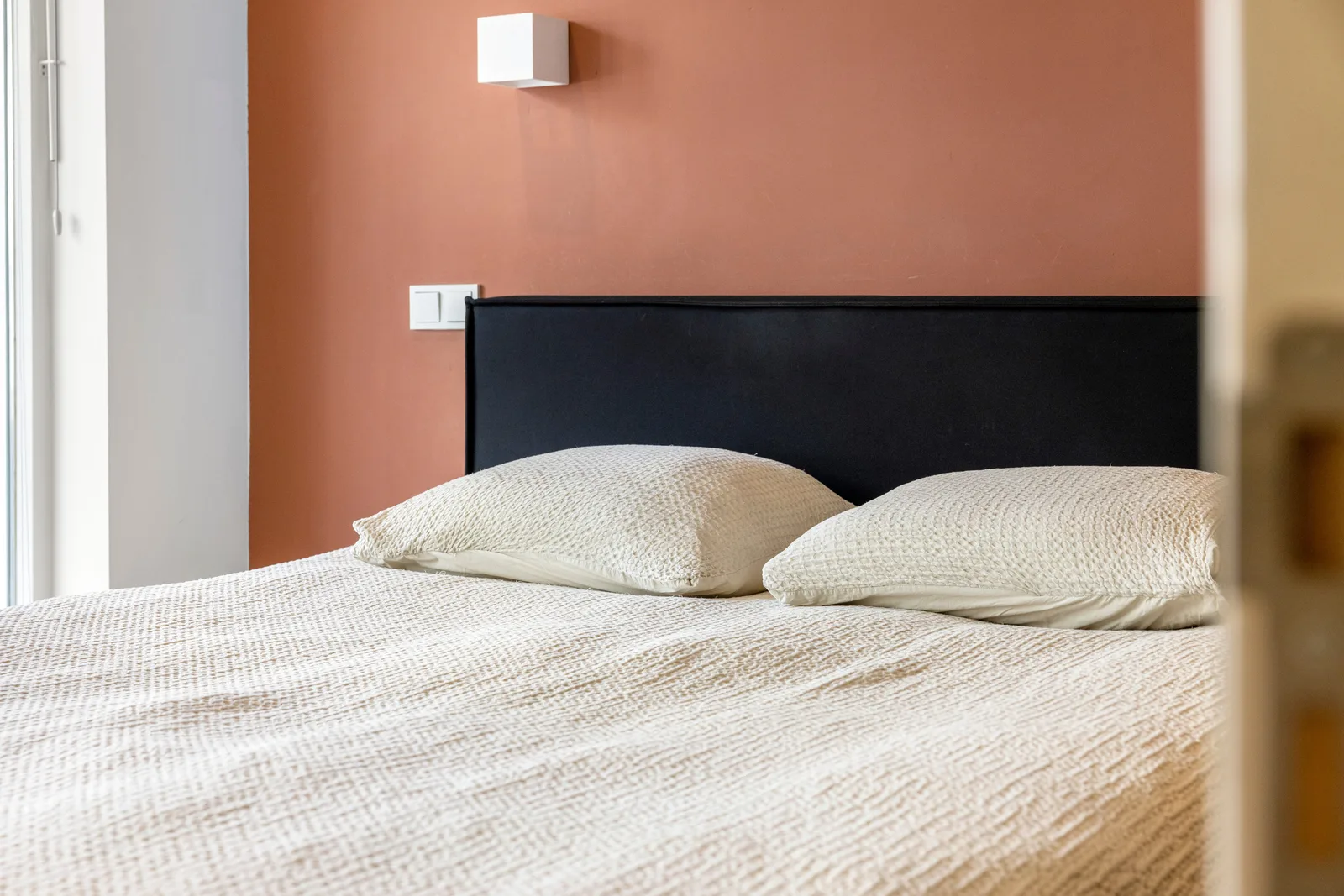Zwart hoofdbord tegen een terracotta wand, met een witte kubuswandlamp en twee crèmekleurige kussens