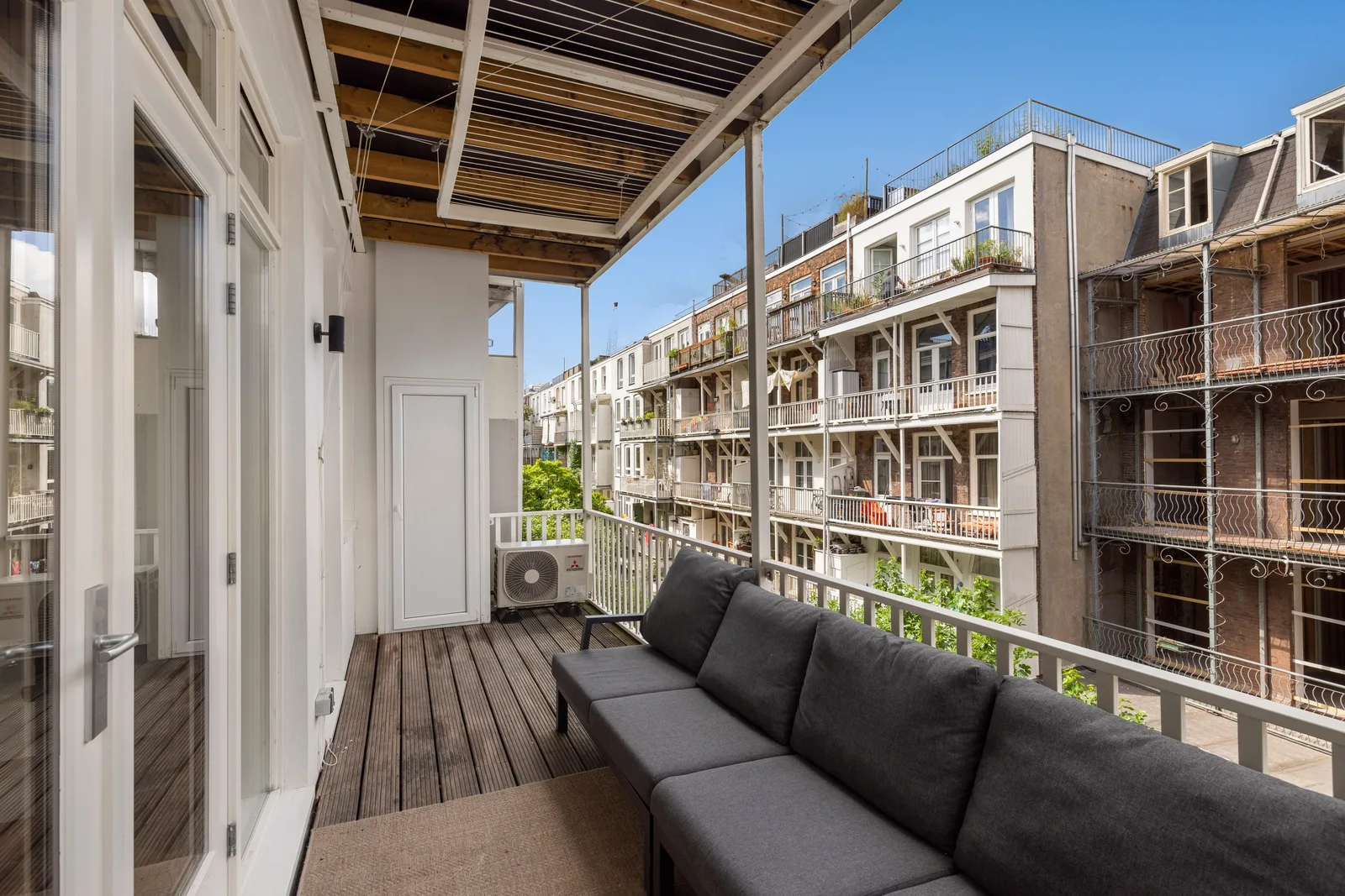 Balkon met houten vlonderdek, donkergrijze hoekbank en een overkapping van houten balken op witte staanders