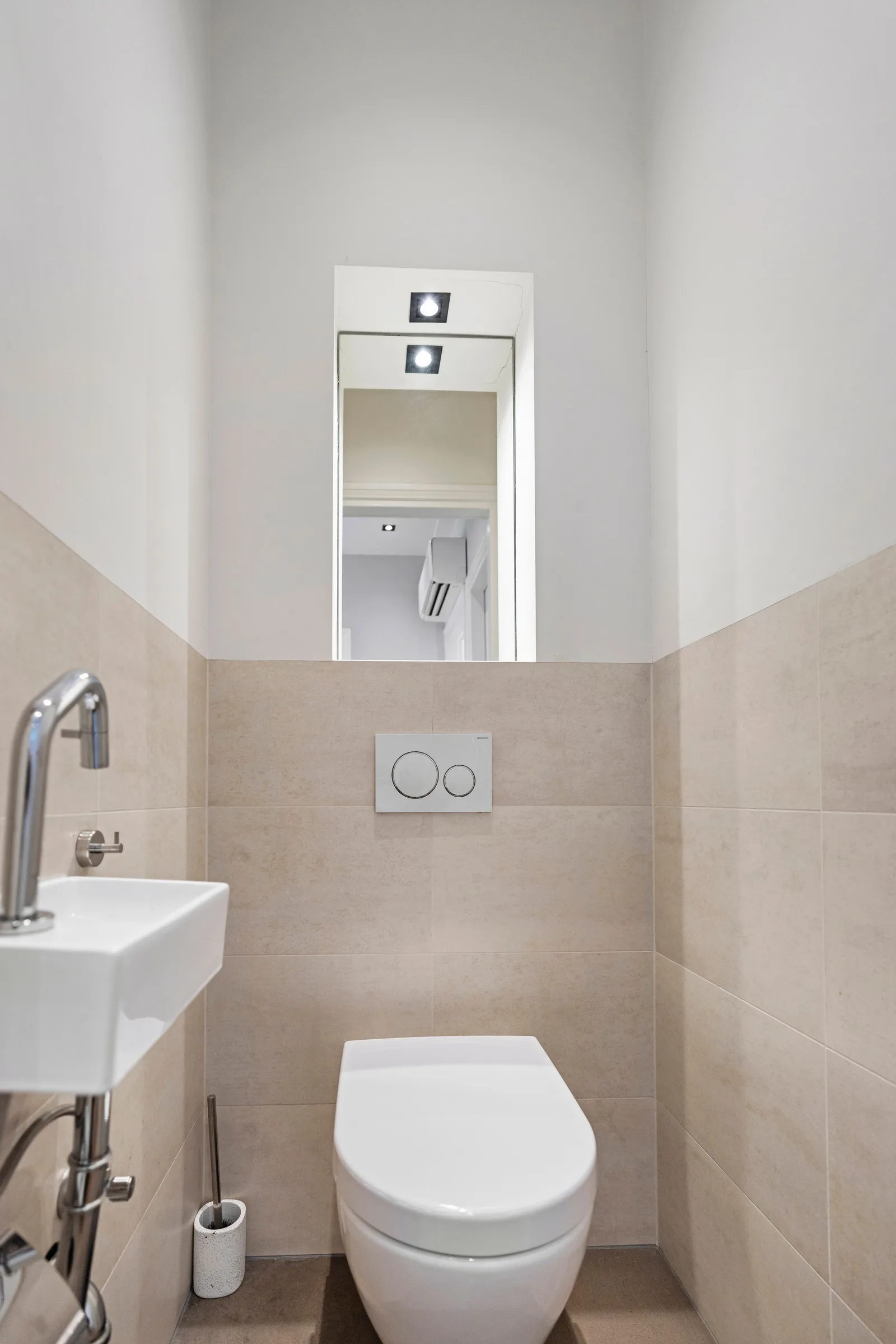 Smal toilet, beige tegels tot halve wandhoogte, wandcloset met spiegel erboven, wit fonteintje met chromen kraan