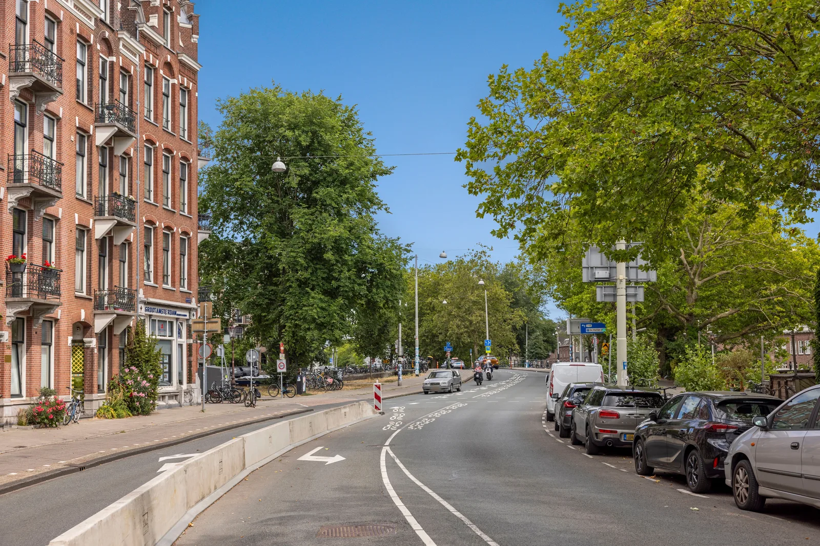 Brede asfaltstraat met lage betonnen rijbaanscheiding, fietsen op de stoep en hoge bomen langs beide kanten