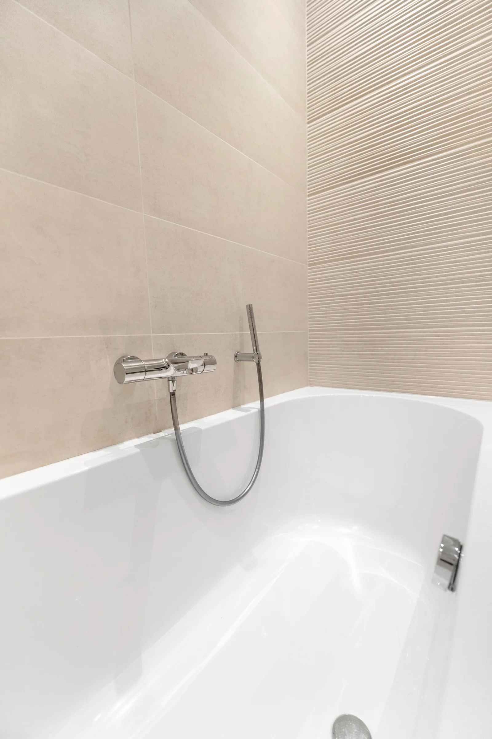 Chroomkleurige badkraan met handdouche boven een wit ligbad, rechts beige reliëftegels