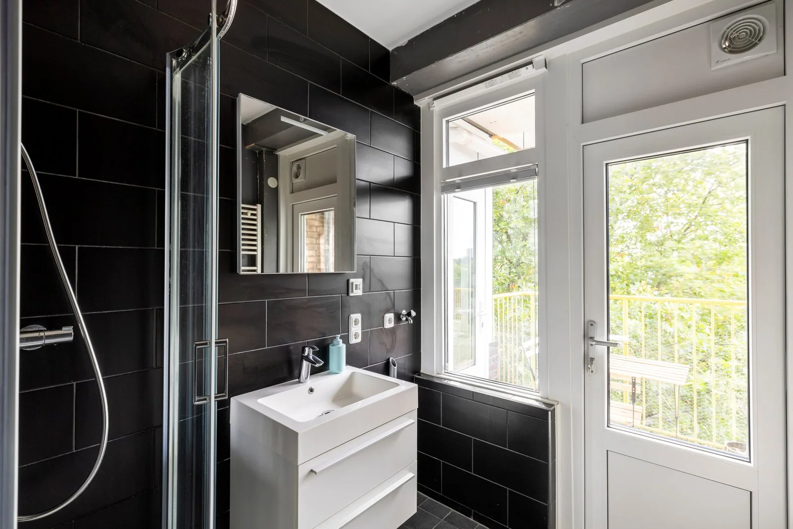 Badkamer met zwarte wandtegels, hoekdouche en wastafelmeubel naast de balkondeur