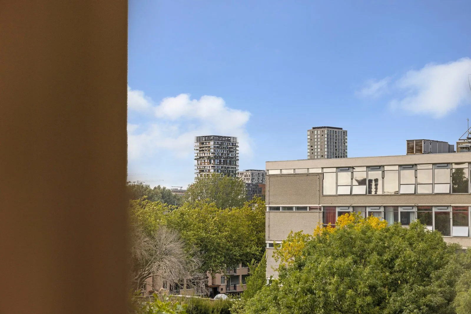 Uitzicht over boomtoppen op een laag gebouw met raamstroken en woontorens in de verte