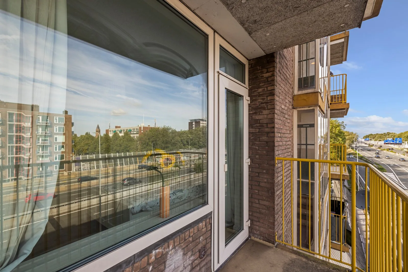 Balkon langs de bakstenen gevel, met geel hek, brede raampartij en balkondeur