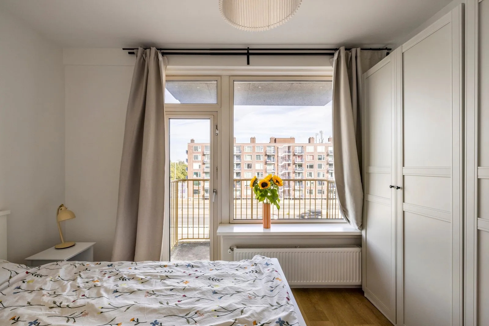 Slaapkamer met balkondeur en raam, uitkijkend op een bakstenen woonblok met balkons