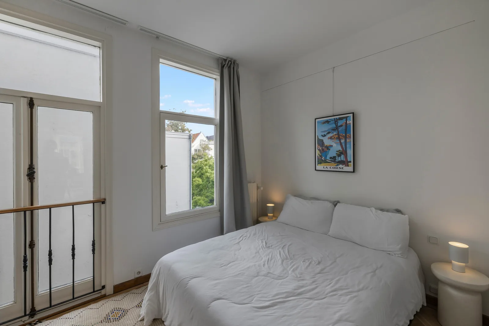 Tweepersoonsbed onder een hoog raam, met een openslaande balkondeur en zicht op boomtoppen