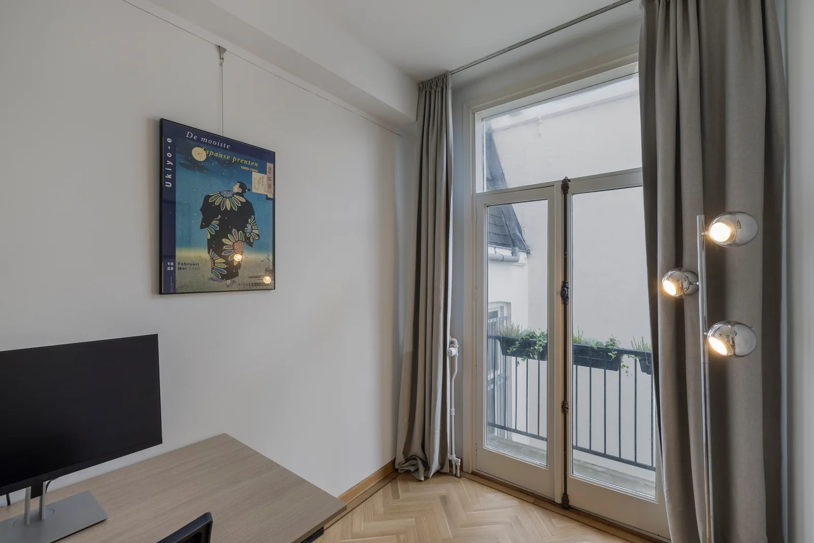 Houten bureau met monitor naast openslaande deuren, op het balkon staan plantenbakken