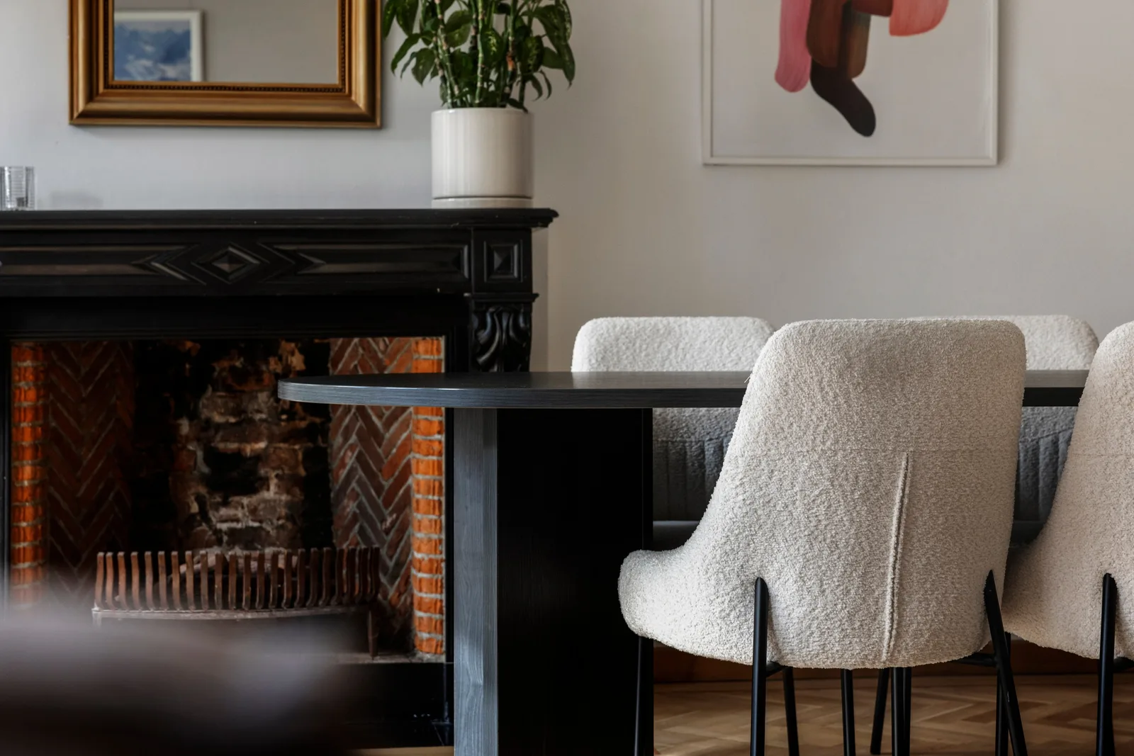 Zwarte houten schouw met snijwerk, haardvloer van rode baksteen, rechts drie bouclé stoelen