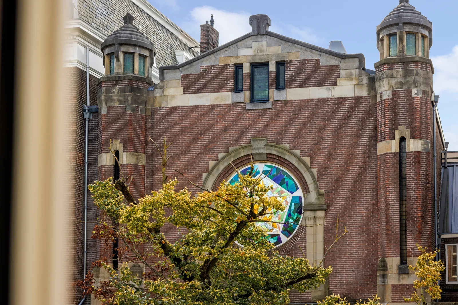 Door de boomkruin de bakstenen kerkgevel, met glas in lood roosvenster en twee hoektorentjes