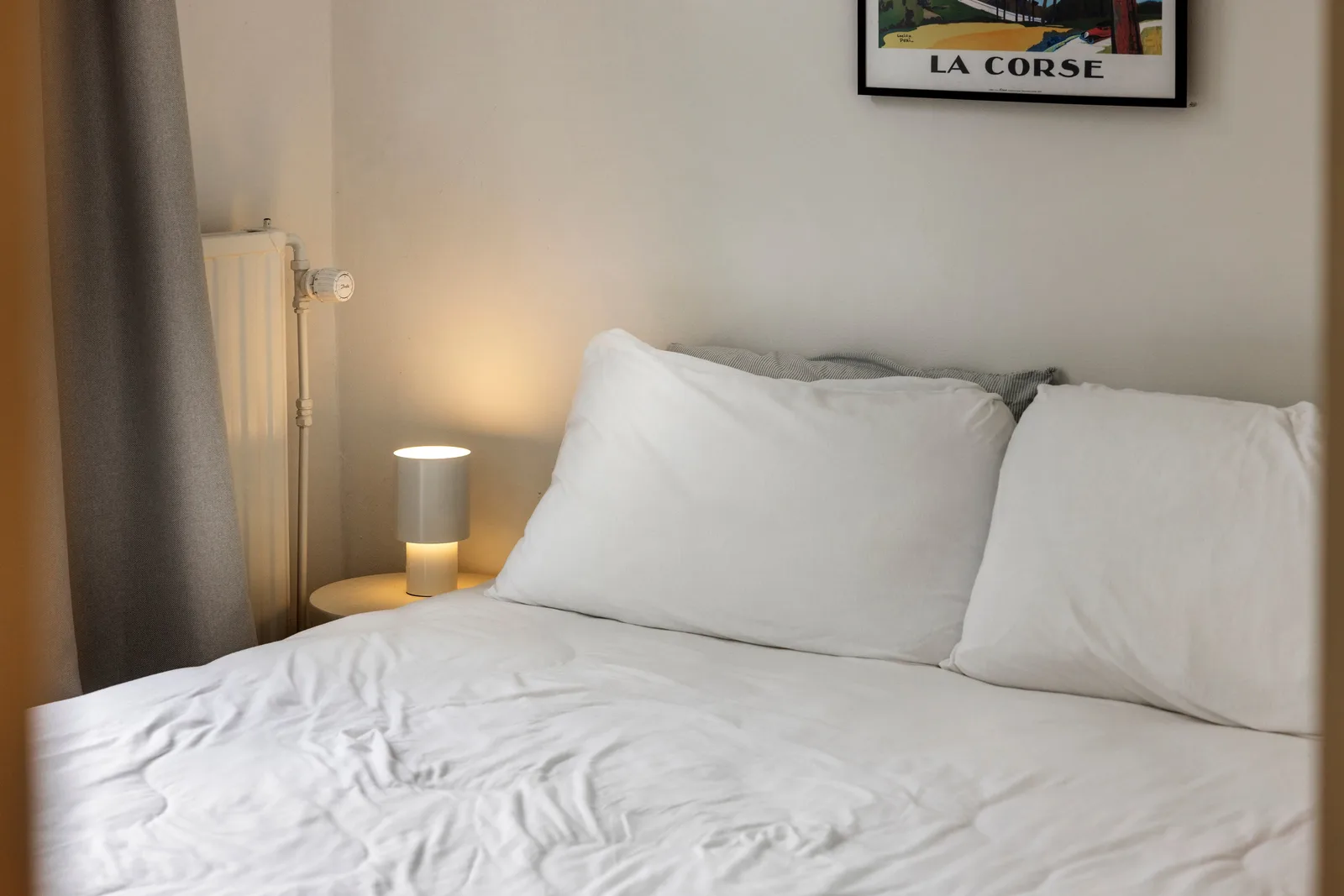 Wit opgemaakt bed tegen de wand, ernaast een gietijzeren radiator en een cilinderlamp