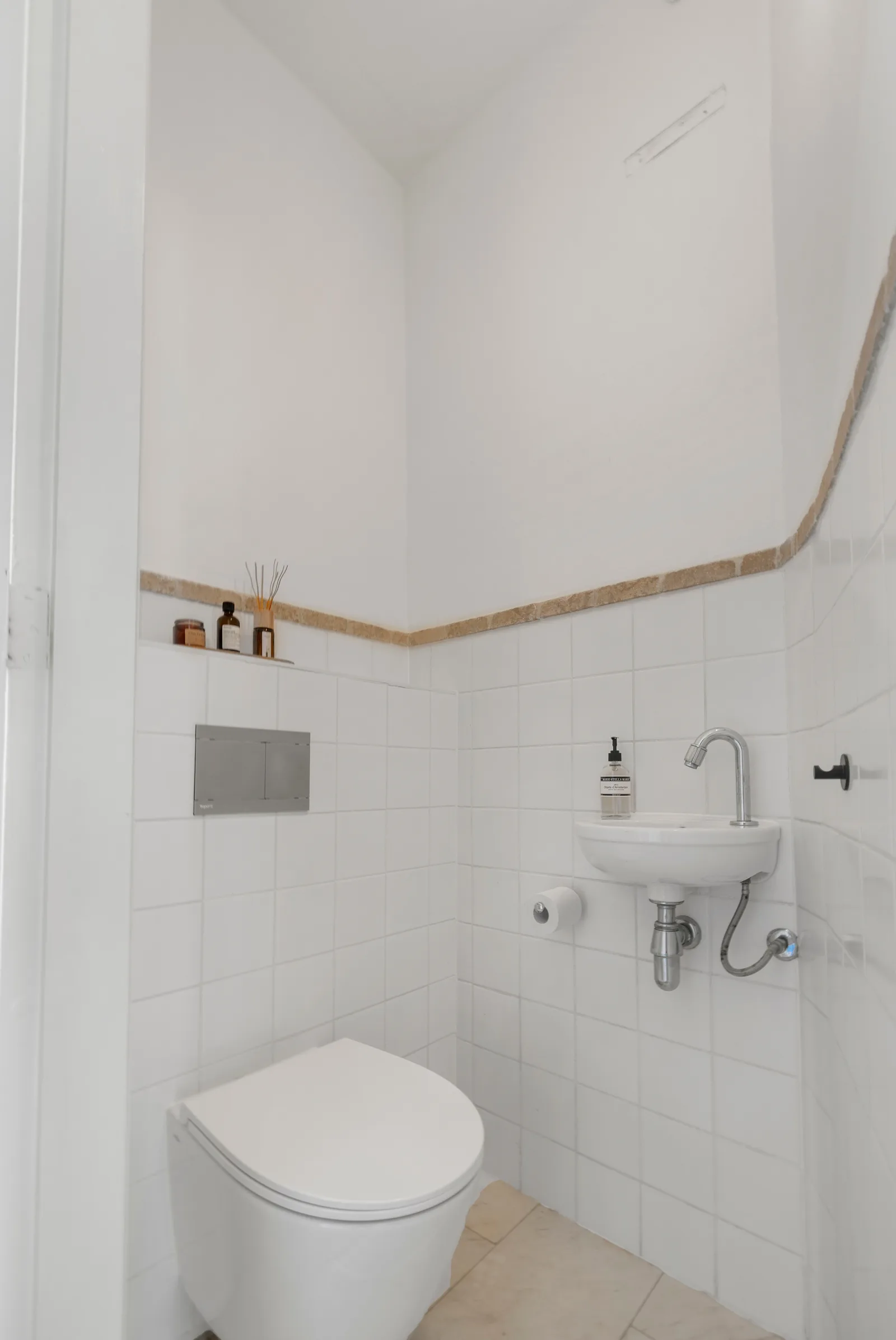 Hangend closet en kleine fontein tegen wit tegelwerk, met beige natuurstenen randstrook