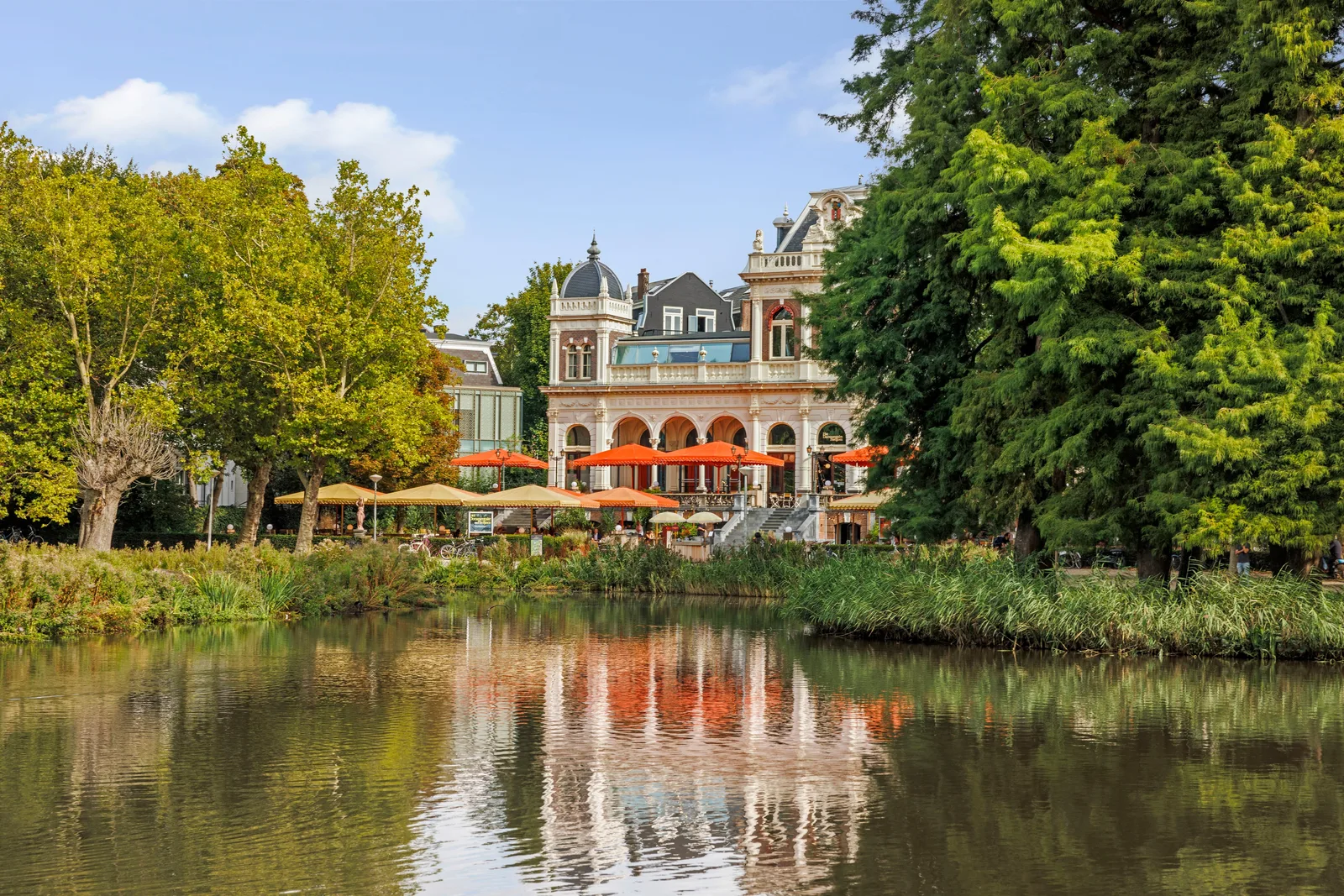 Het paviljoen spiegelt in het water, op de voorgrond riet en gekleurde parasols