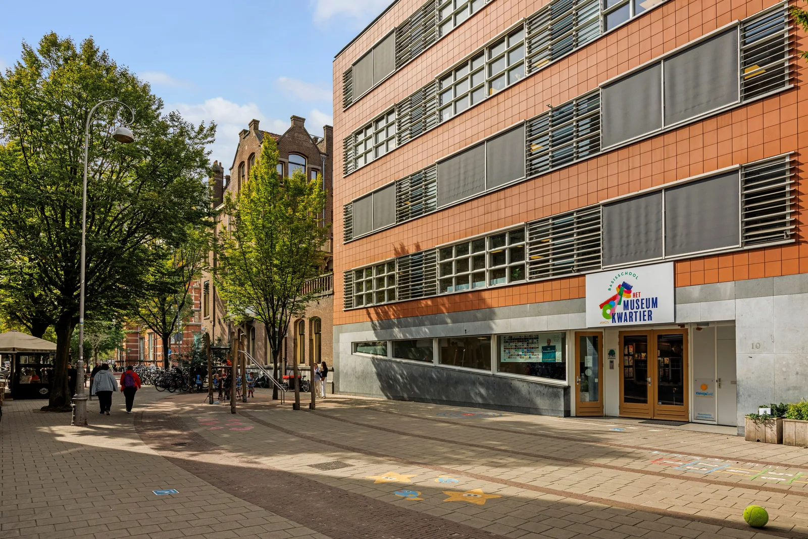 Basisschool met terracottakleurige gevelpanelen, speelfiguren in de bestrating en bomen langs de stoep