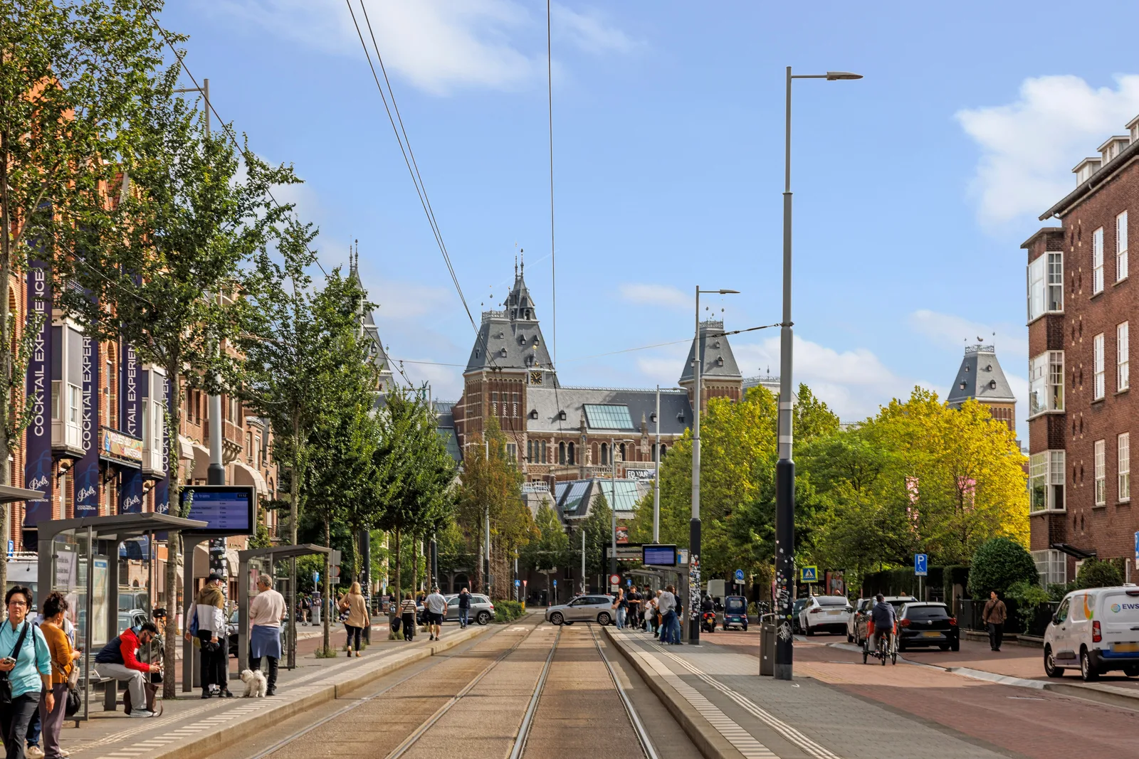 Bij de tramhalte wachten mensen, aan het eind van de straat staat het Rijksmuseum