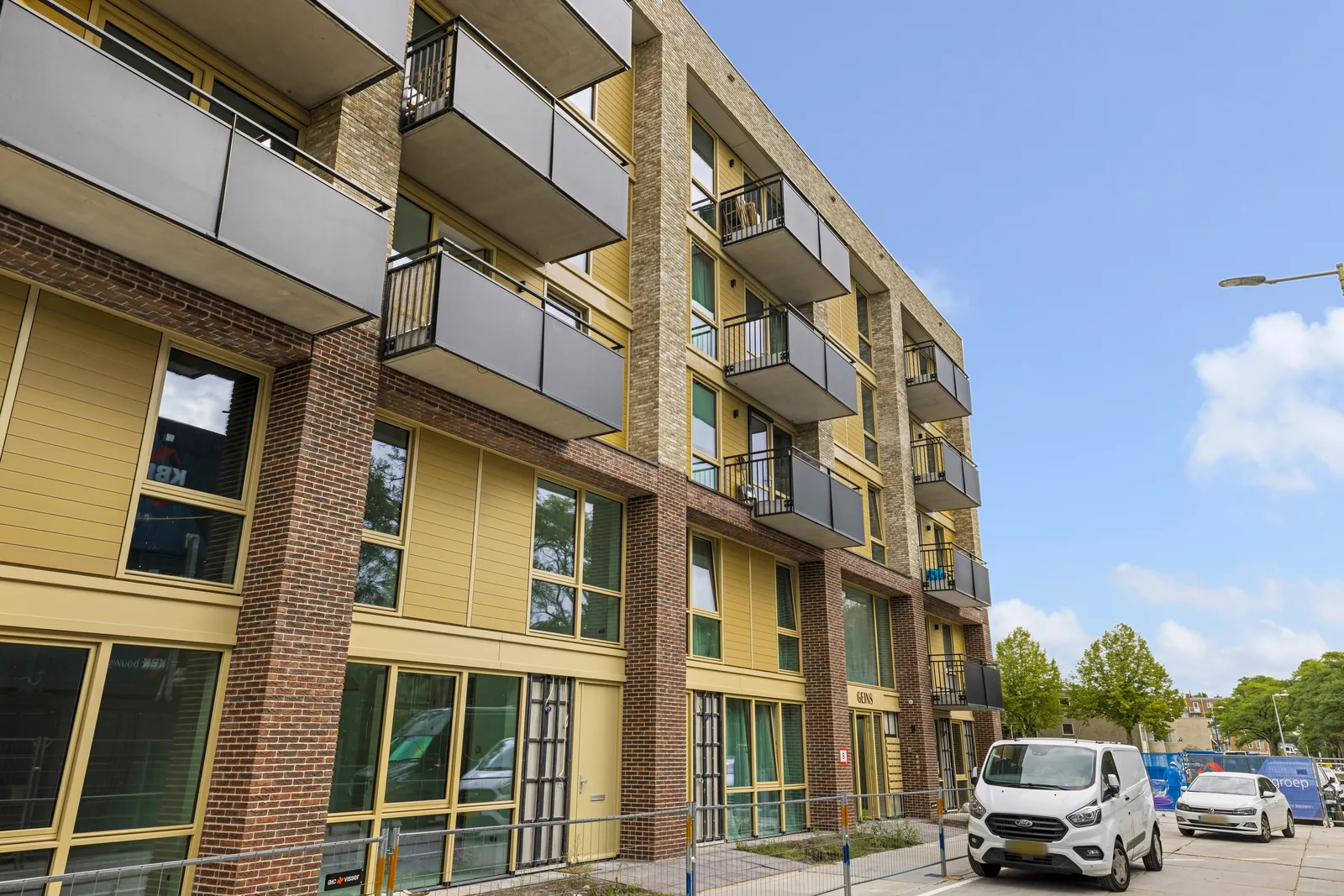 Appartementengebouw met gele gevelpanelen, roodbruin metselwerk en lichtgrijze balkonpanelen, ervoor een witte bestelbus