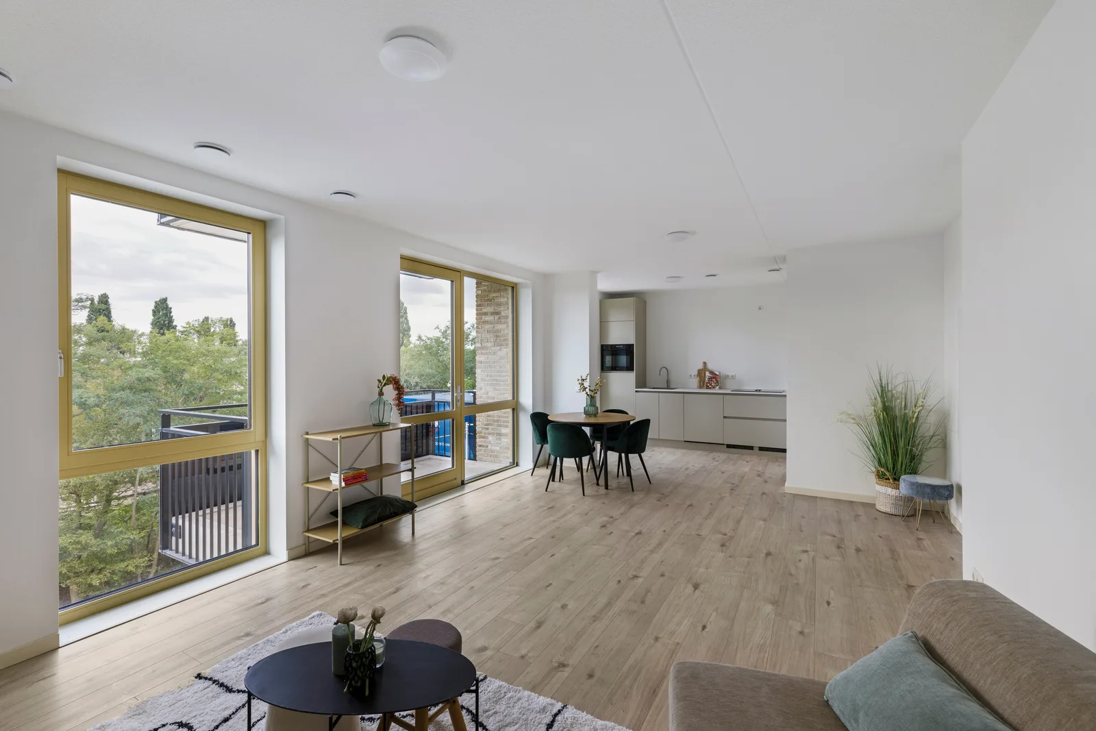 Woonkamer met lichte plankenvloer en okergele kozijnen, achterin een lichtgrijze keuken en ronde eettafel