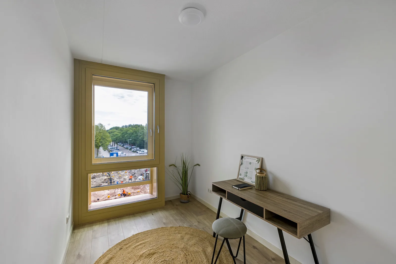 Kleine kamer met okergeel raamkozijn tot bij de vloer, houtlook bureau, kruk en ronde jute mat