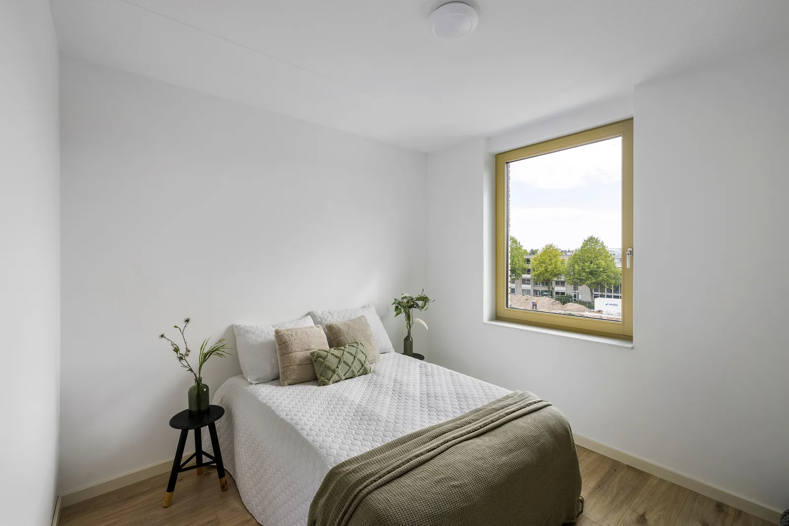 Slaapkamer met een tweepersoonsbed, witte sprei en groene deken, rechts een okergeel raamkozijn