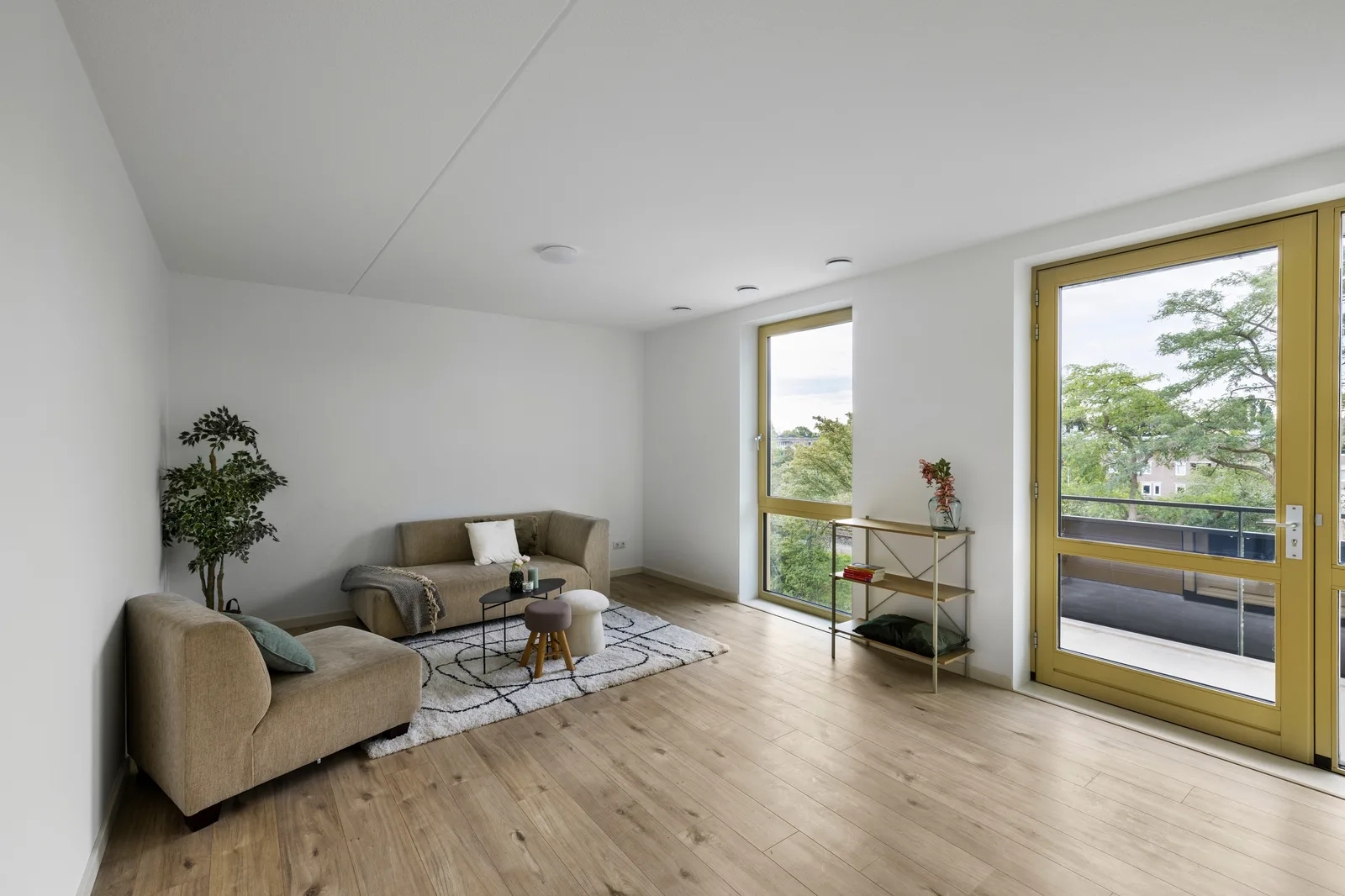 Woonkamer met een beige bank en chaise longue, lichte houten vloer en okergele balkondeur