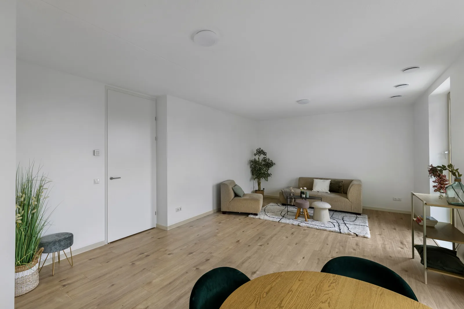 Woonkamer met witte wanden en lichte houten vloer, beige bank achterin, ronde eettafel met donkergroene stoelen