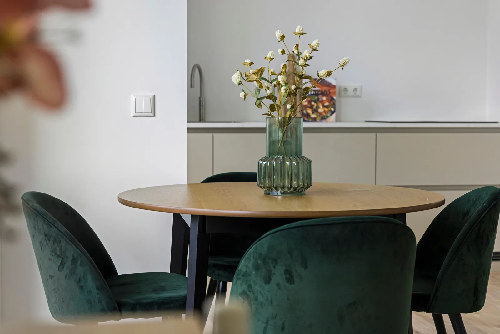 Ronde houten eettafel met donkergroene velours stoelen en een groene glazen vaas met droogbloemen