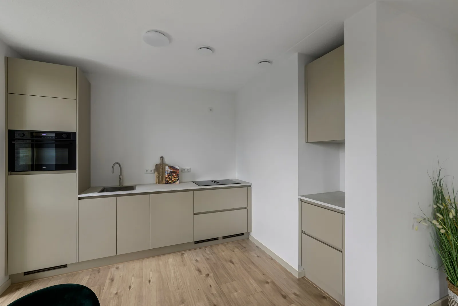 Zandkleurige greeploze keuken met spoelbak en kookplaat in een licht blad, links een hoge ovenkast