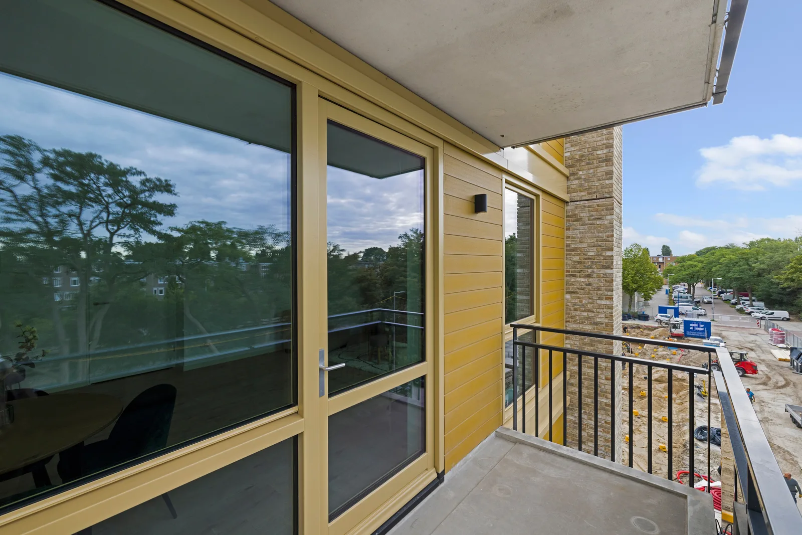 Balkon met glazen pui, okergele gevelbekleding en zwart spijlenhek, rechts zicht op een bouwterrein