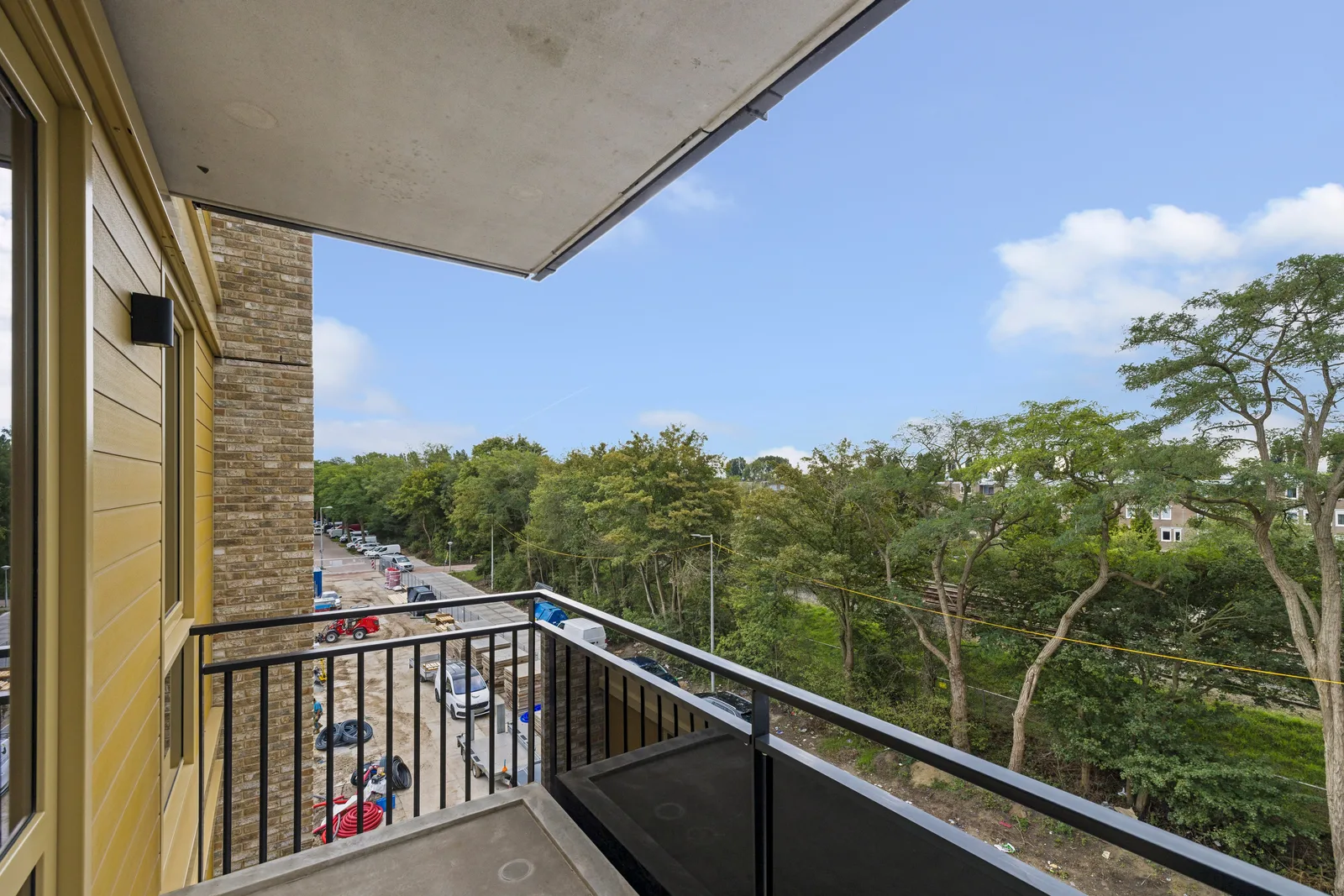Uitzicht vanaf het balkon op hoge bomen en woningen, beneden een straat en een bouwterrein