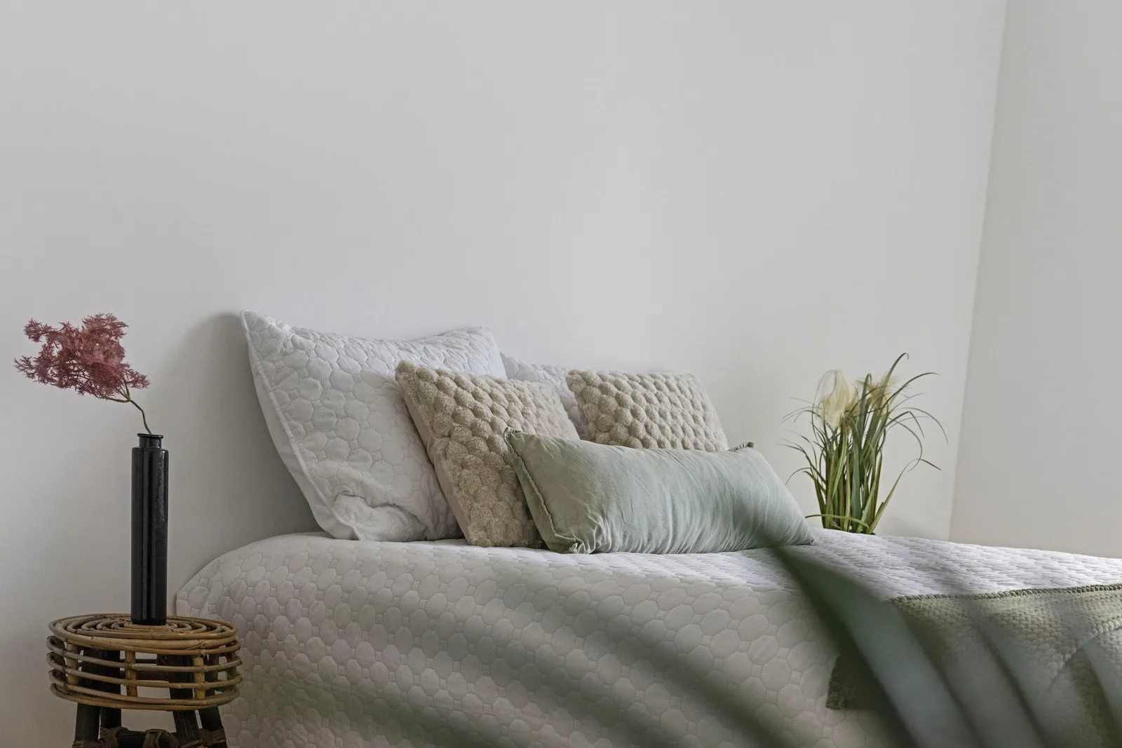 Bed met witte gewatteerde sprei, beige kussens en groene plaid, links zwarte vaas op rieten kruk