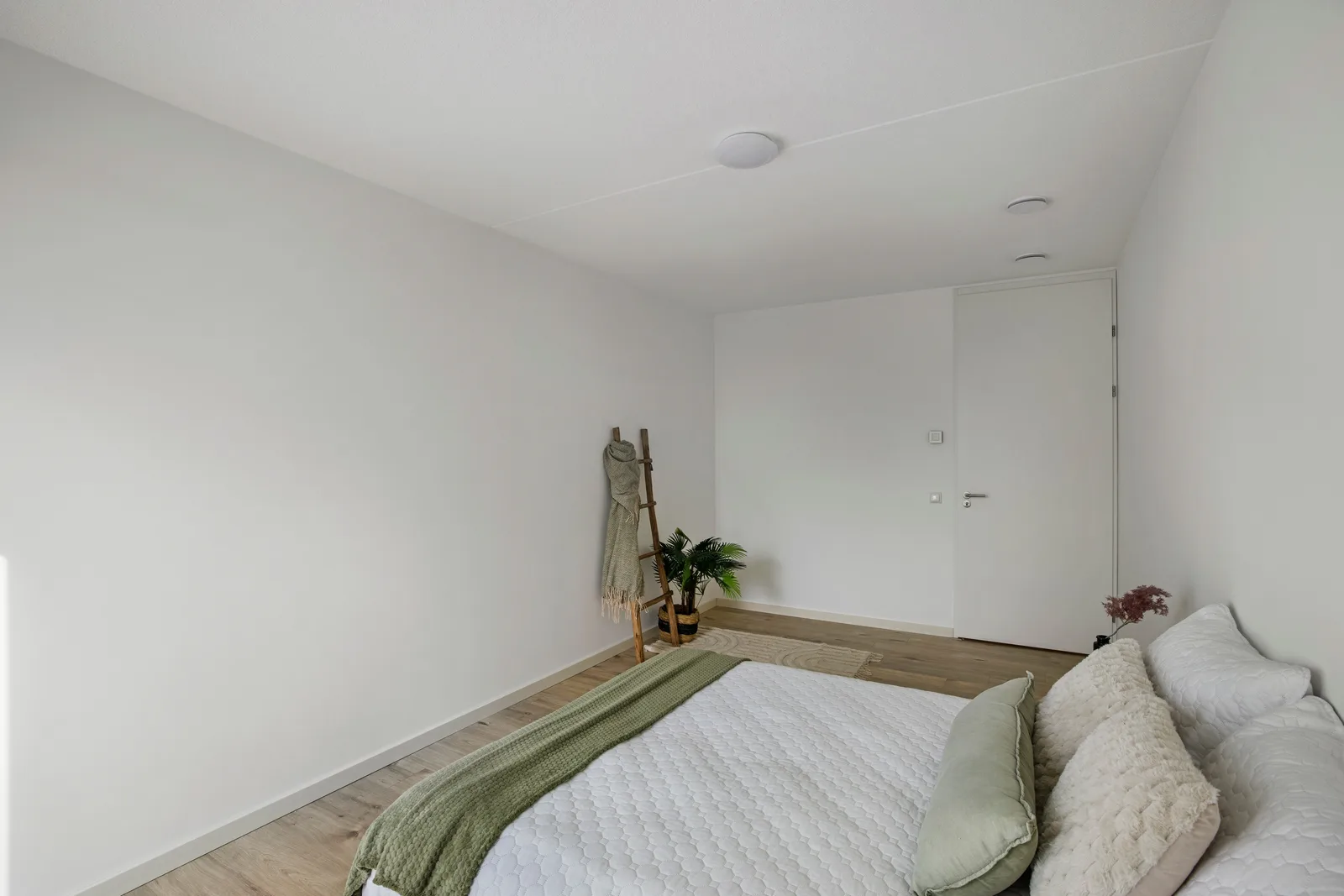 Slaapkamer met wit dekbed en groene plaid, lichte houten vloer, decoratieladder bij de witte binnendeur