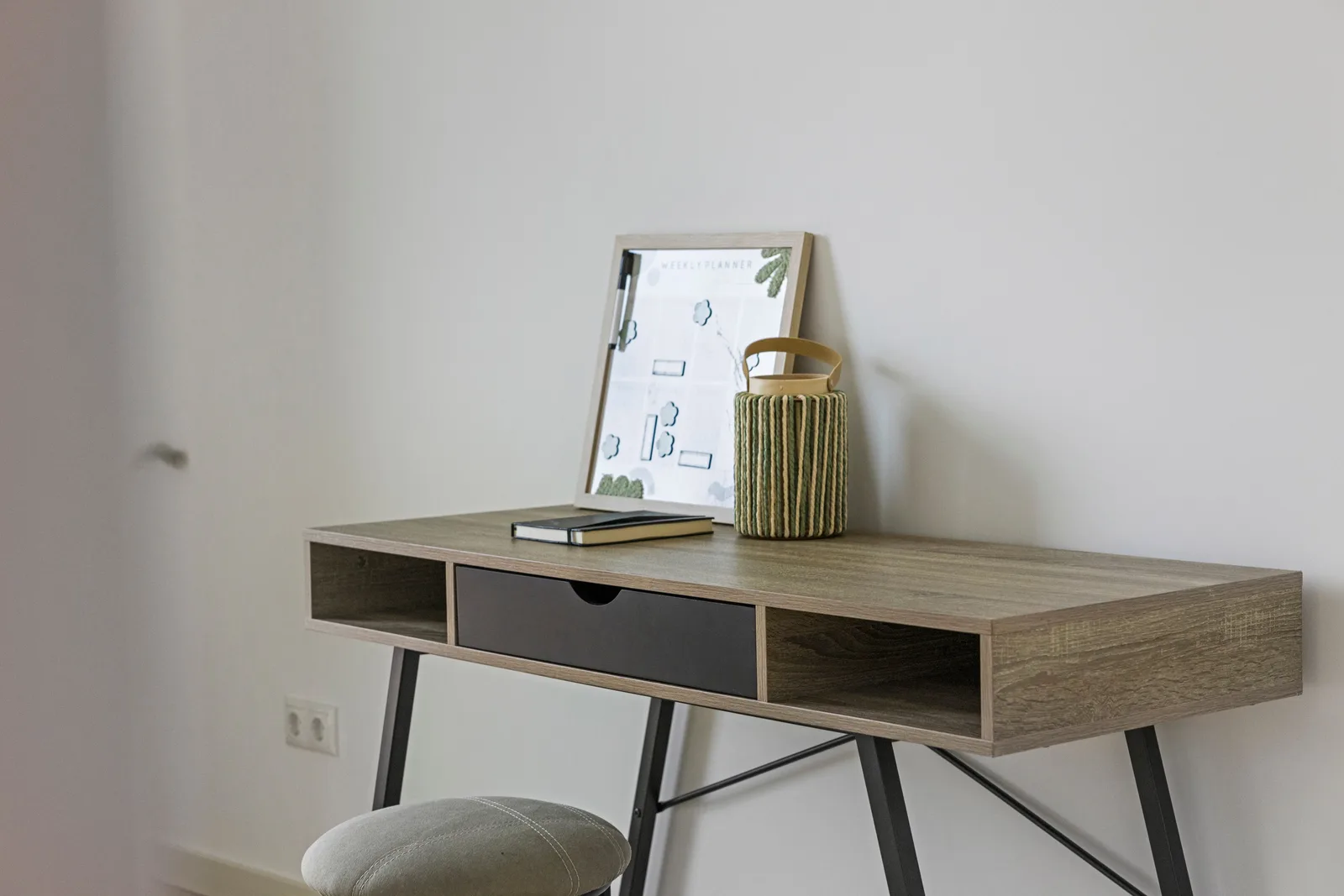 Houtkleurig bureau met zwarte schuine poten, donkere lade en open vakken, ervoor een beige kruk