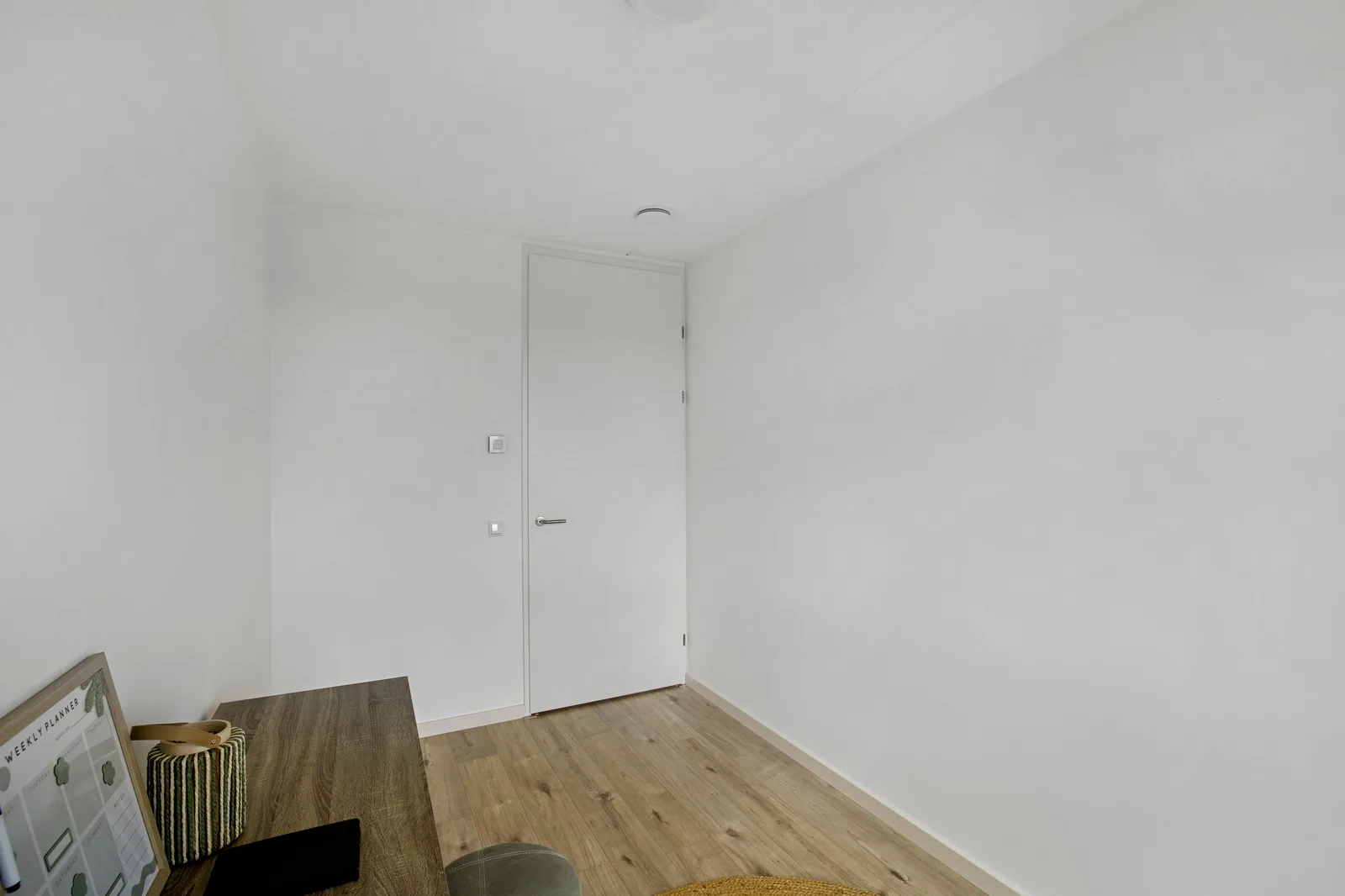 Witte kamer met lichte houten vloer, een witte binnendeur en links een donker houten bureaublad