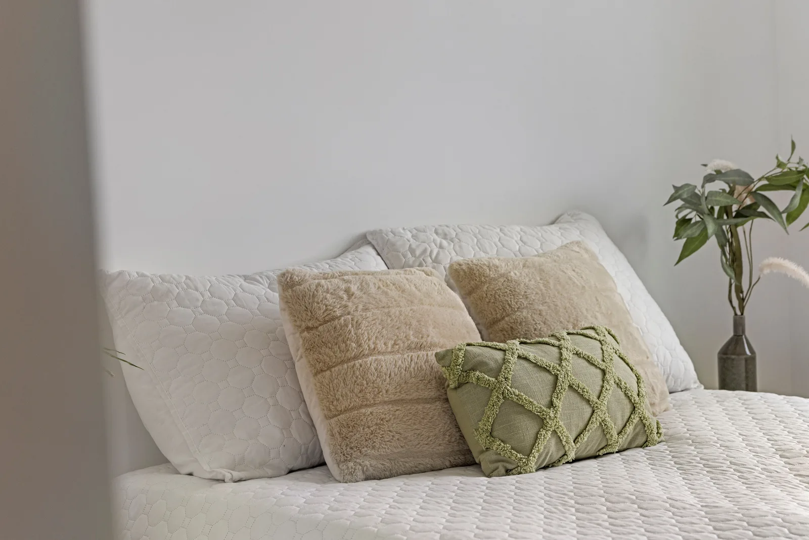 Bed met witte doorgestikte kussens, twee beige en een groen sierkussen, rechts een vaas met takken